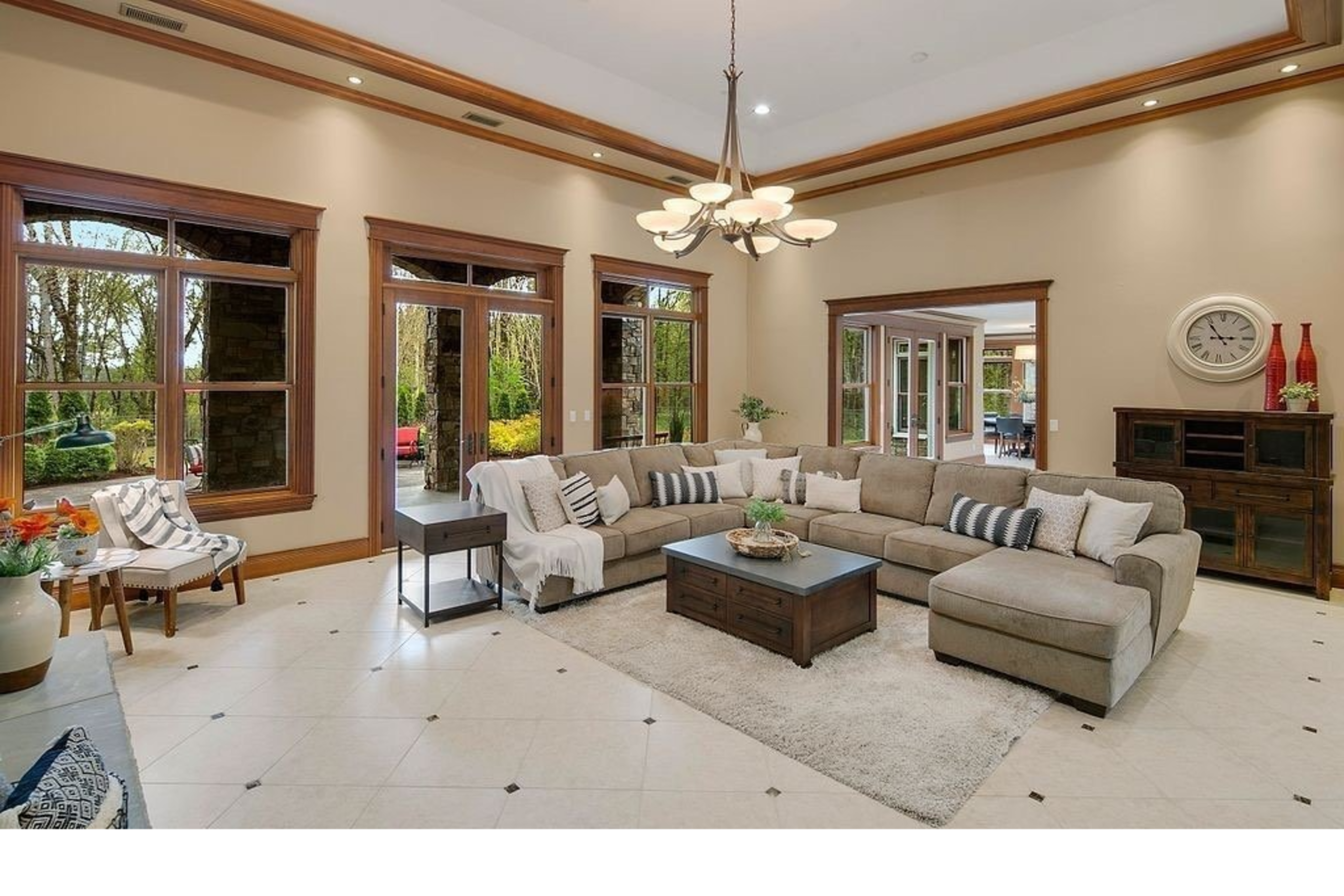 The image shows a spacious living room with a large sectional sofa, a coffee table, and several windows that offer views of greenery outside.