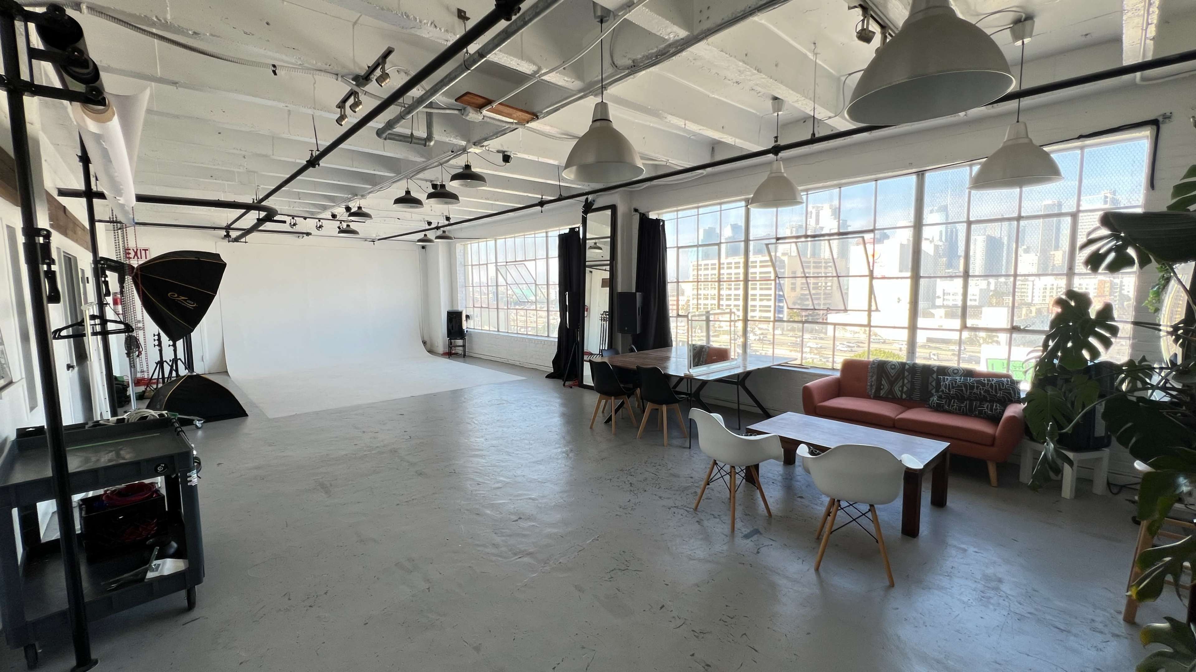 The image shows a spacious, bright studio with large windows, a white backdrop, a table with chairs, and a couch.