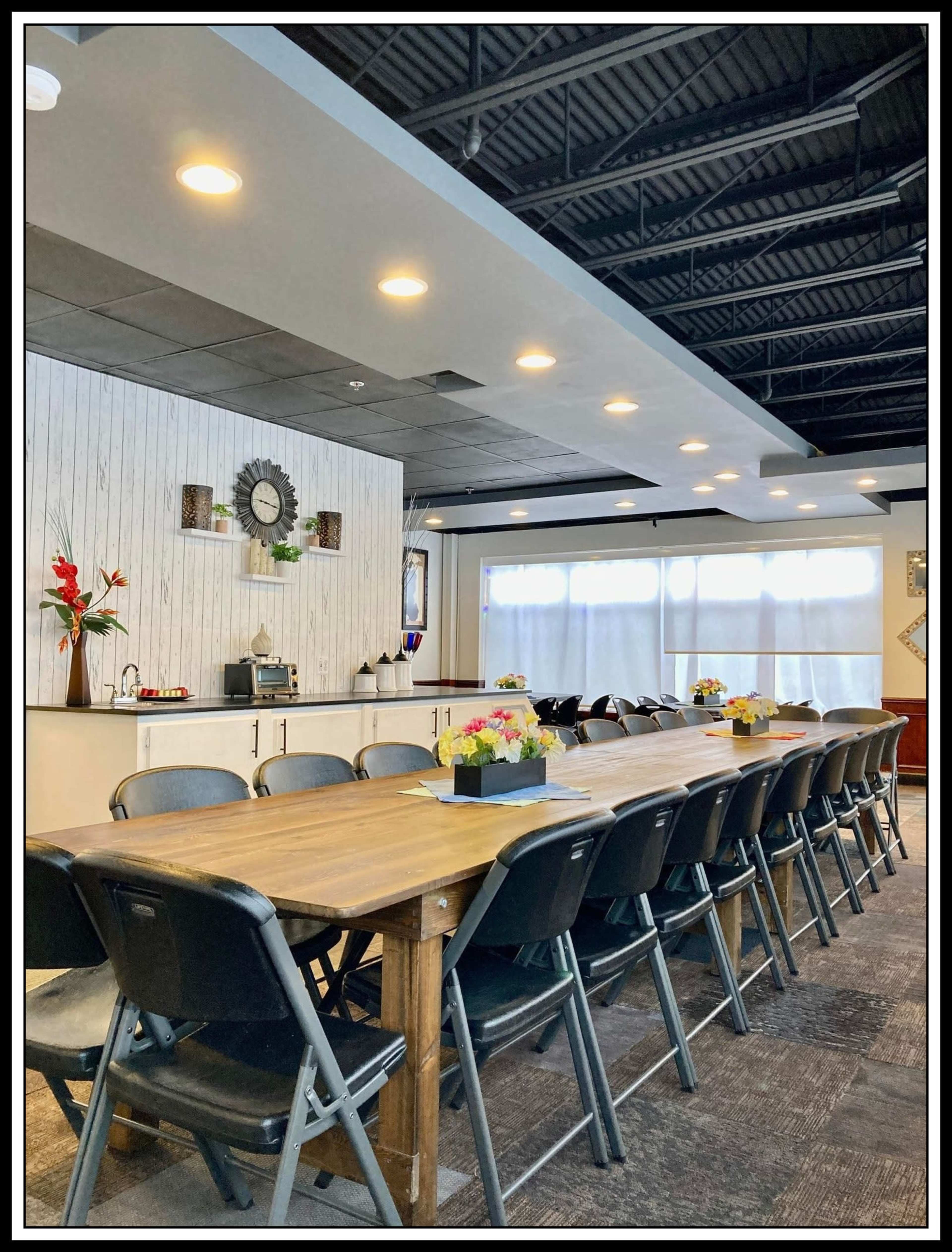 A long wooden table set with flower arrangements in a well-lit room with a kitchenette and large windows.