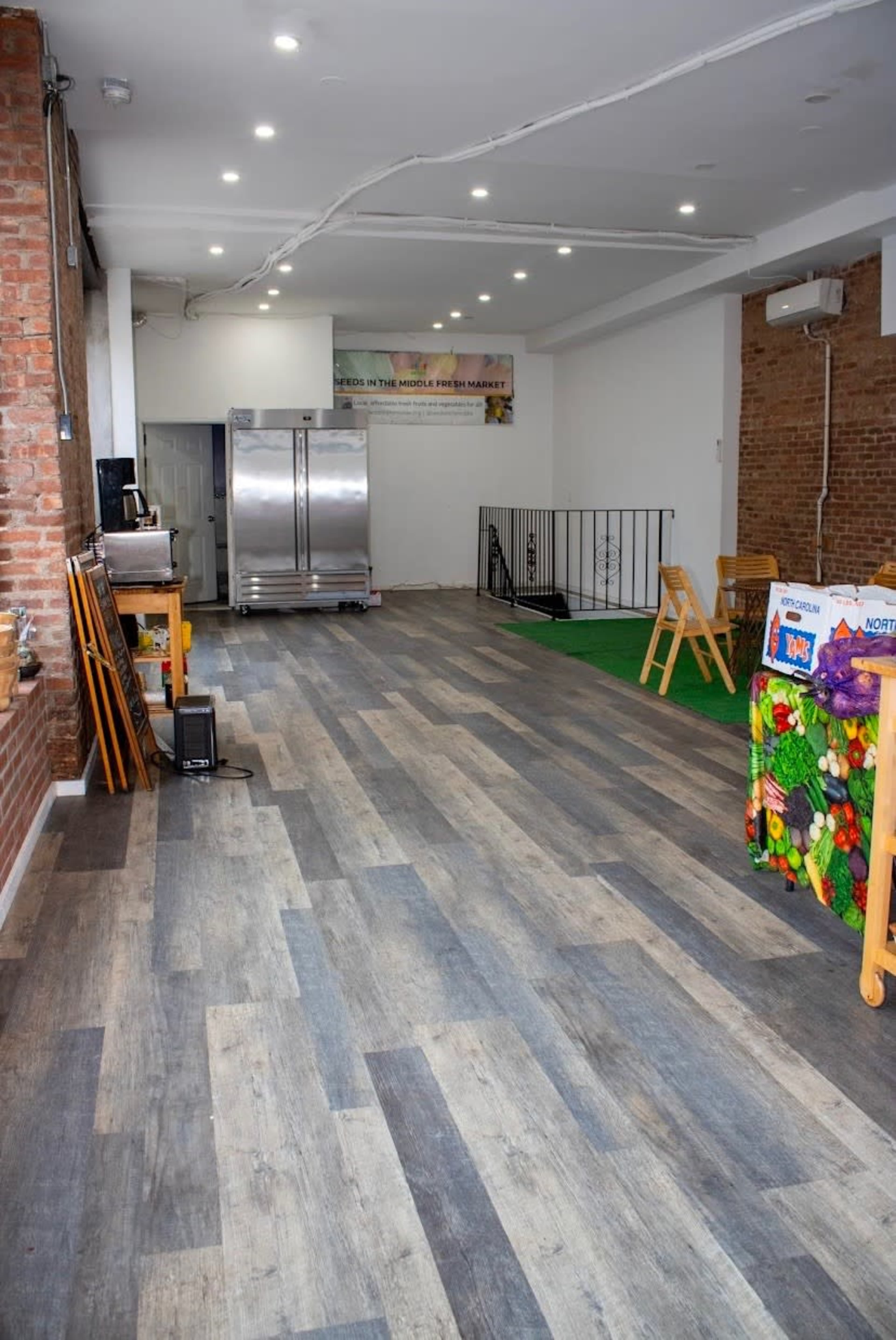 The image shows a spacious interior of a market or store with various materials on the floor and a counter displaying fruits and vegetables in one corner.