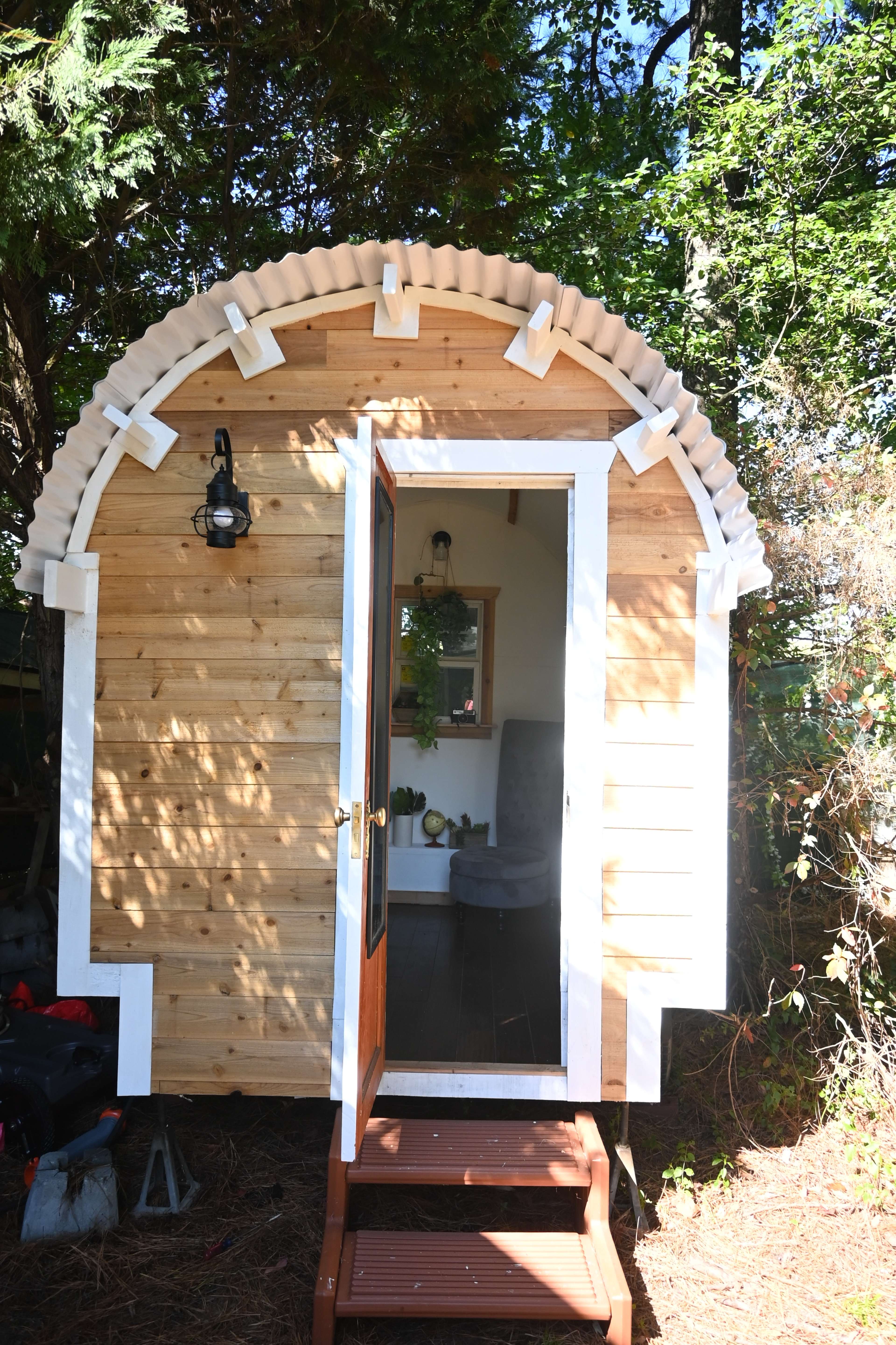 A small, wooden structure with a rounded roof and a front door is surrounded by greenery.