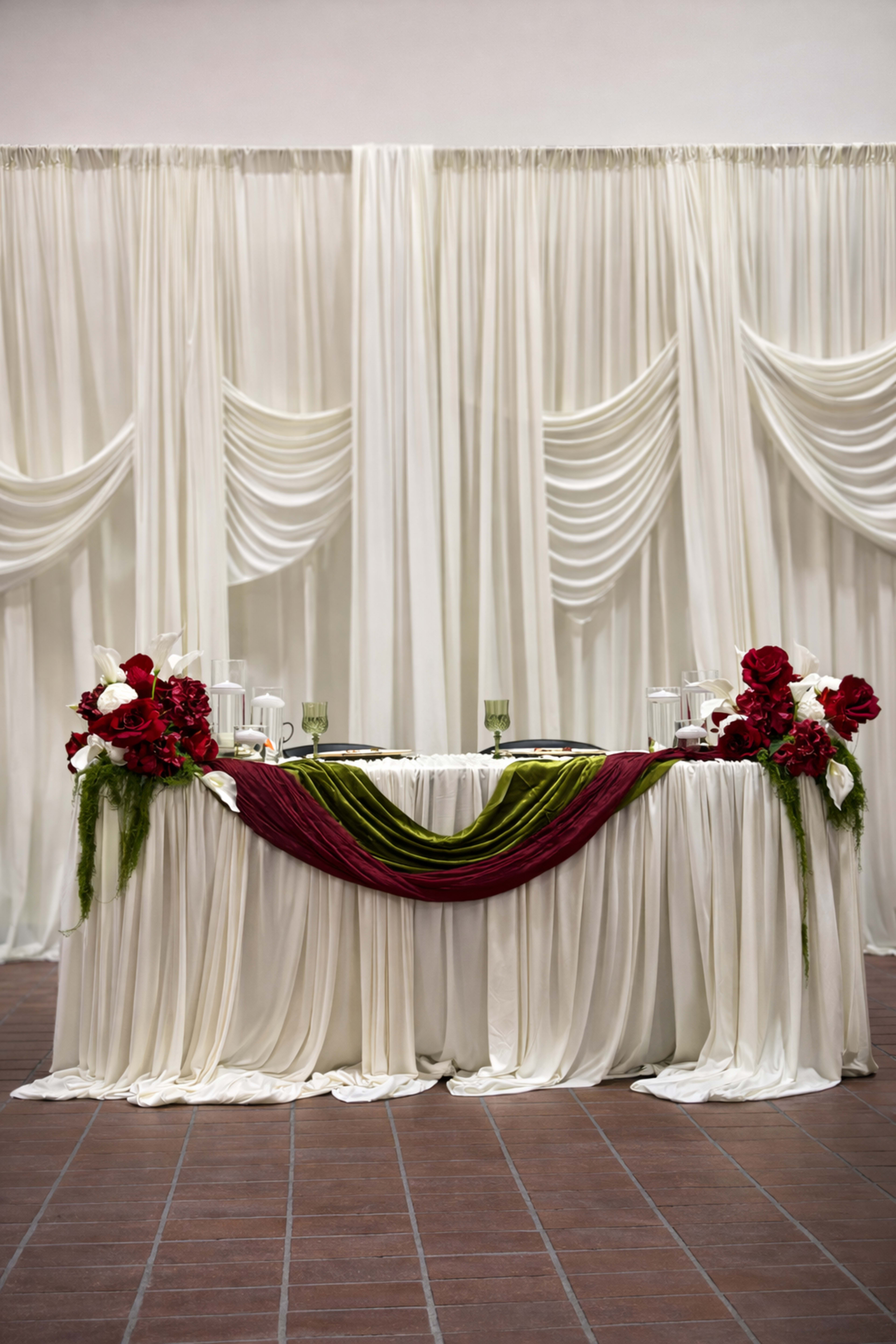 A table is elegantly dressed in white drapery, adorned with red roses and a green scarf, set against a backdrop of similarly draped white fabric.