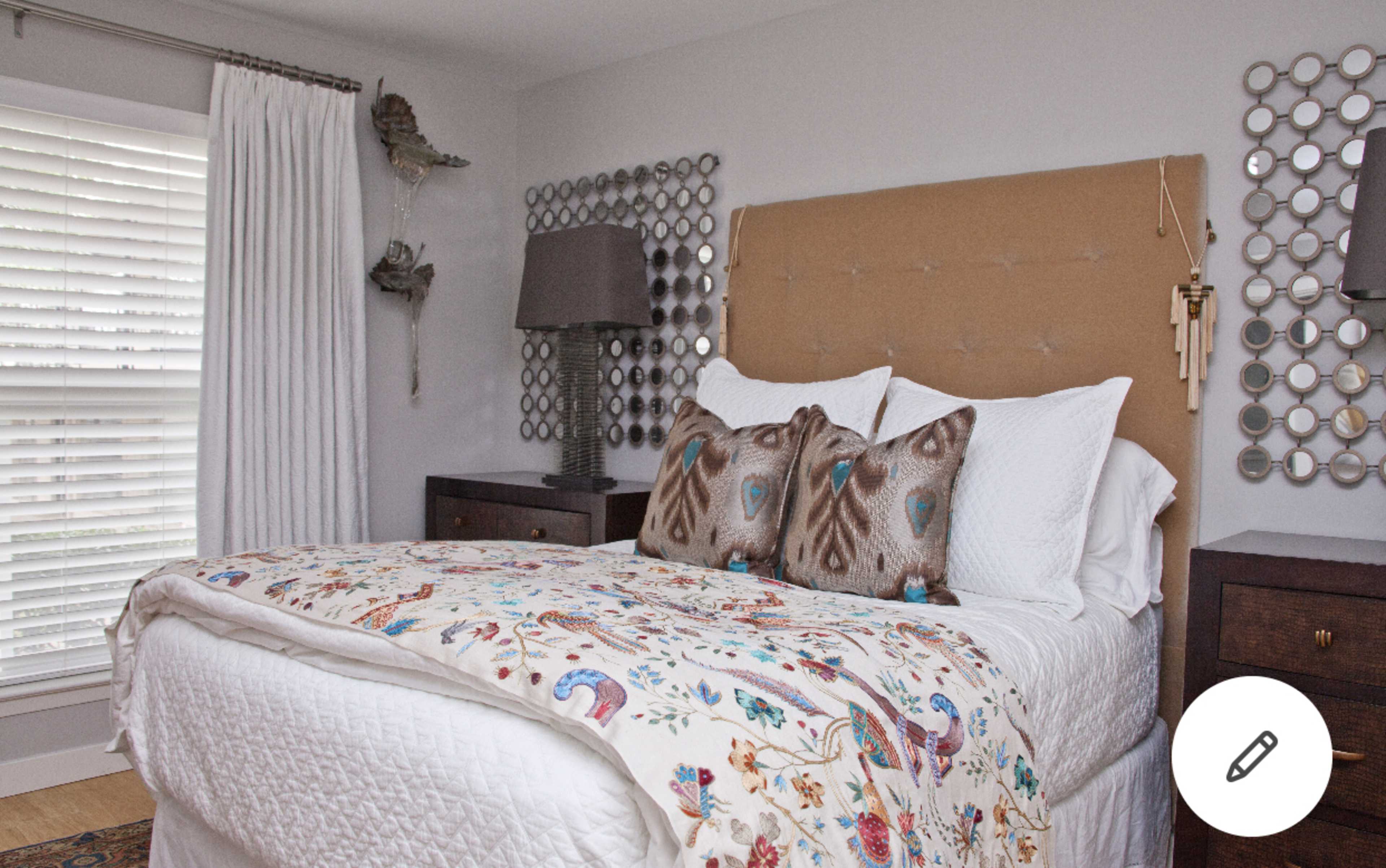 The bedroom features a bed with a patterned quilt and decorative pillows, located against a wall with circular mirrors and a lamp beside it, along with sheer curtains covering a window.