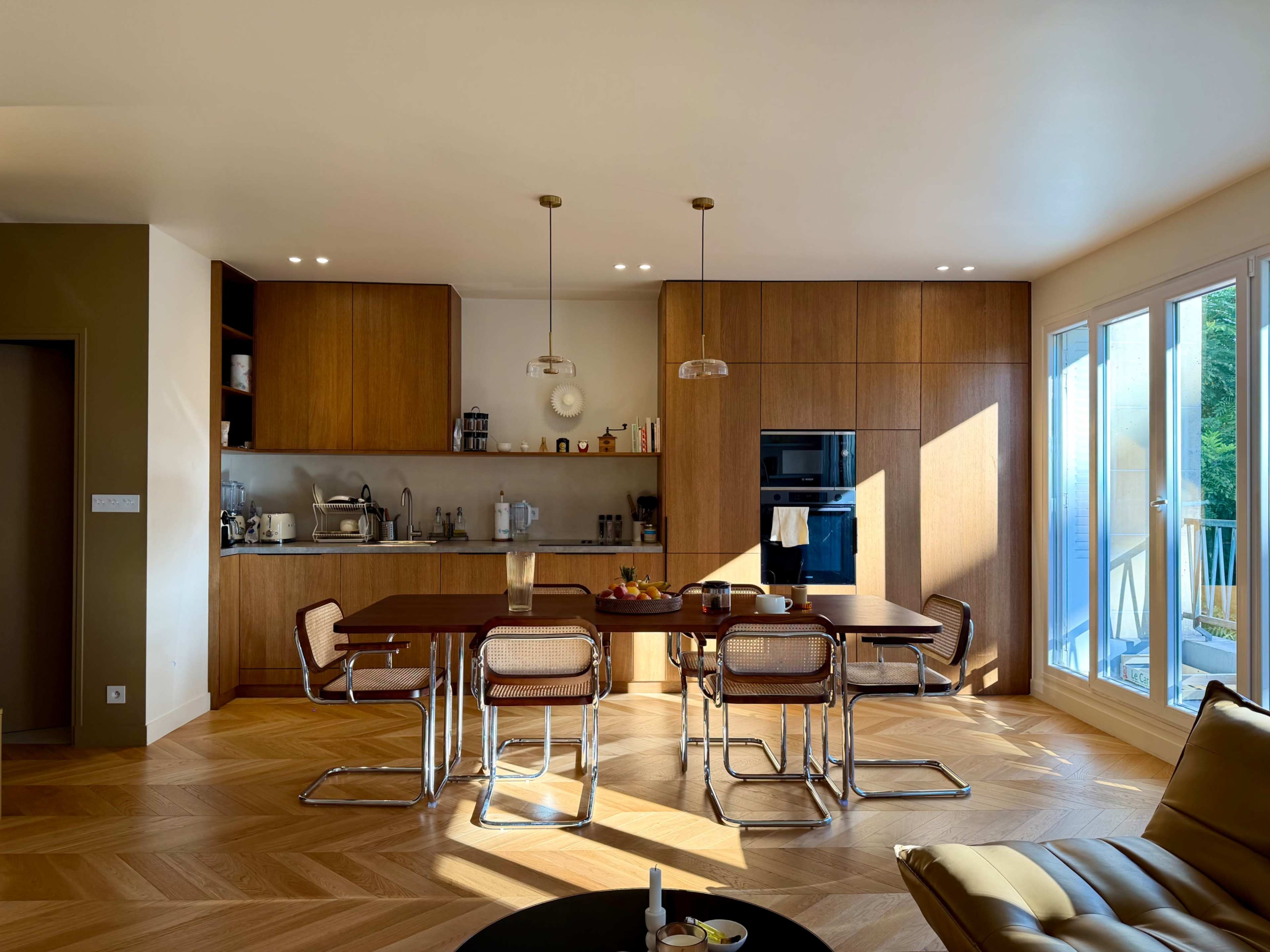 A modern kitchen features wooden cabinetry, a dining table with chairs, and abundant natural light streaming through large windows.