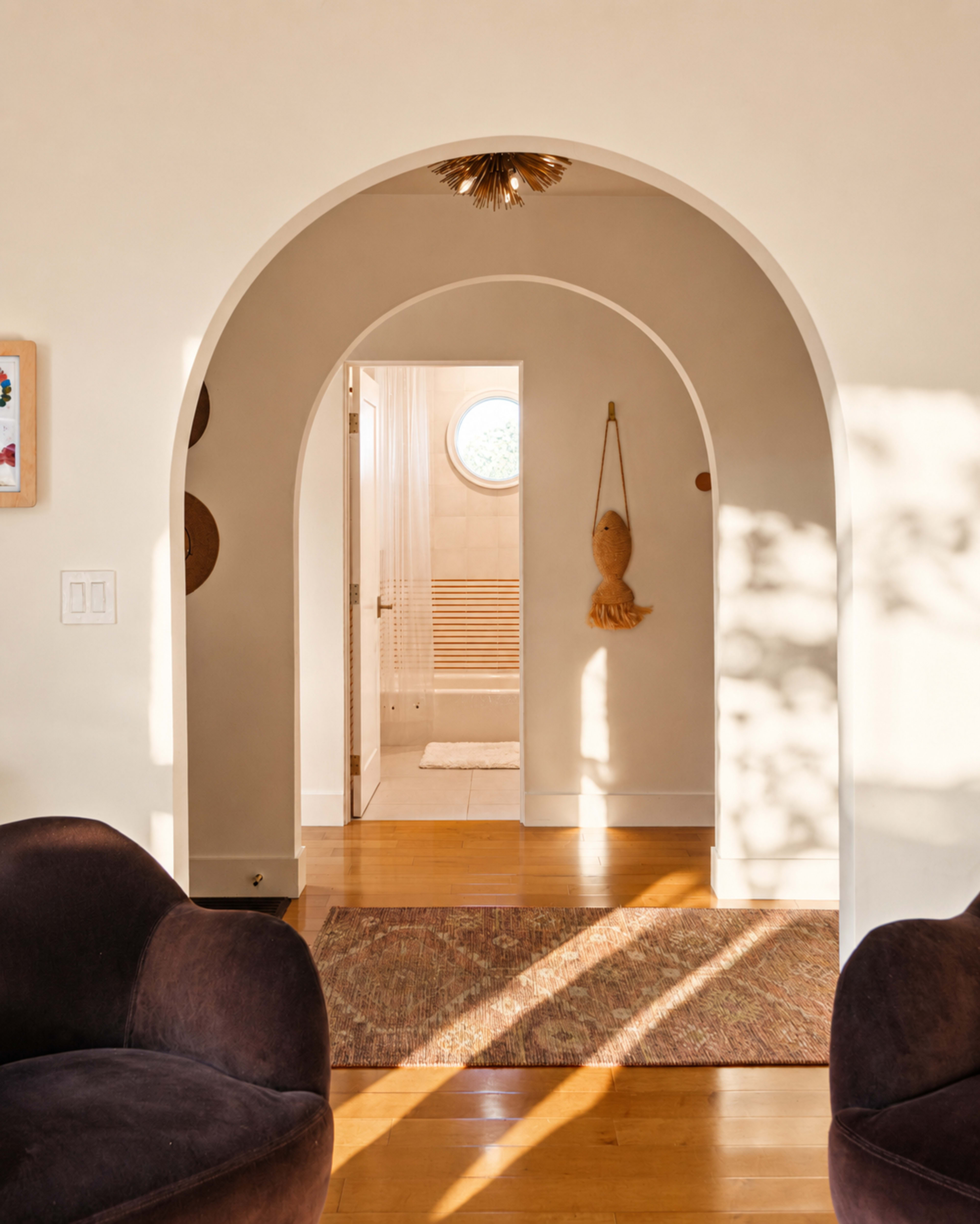 An arched doorway leads to a well-lit bathroom with circular window, framed by a neutral-toned interior featuring two plush chairs and a textured rug.