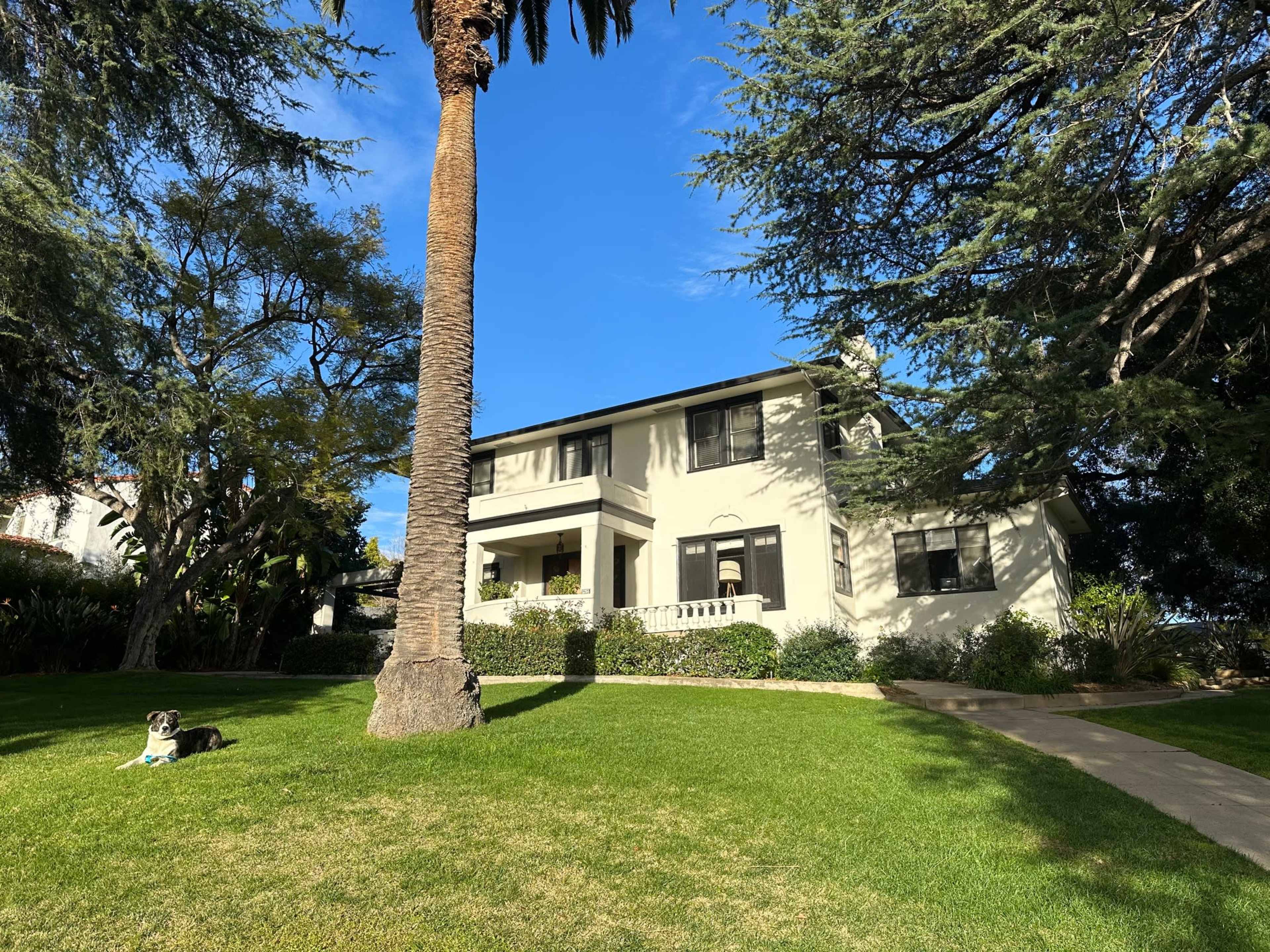 A two-story house with a light-colored exterior sits amidst grassy grounds and tall trees, with a dog lying on the lawn in the foreground.