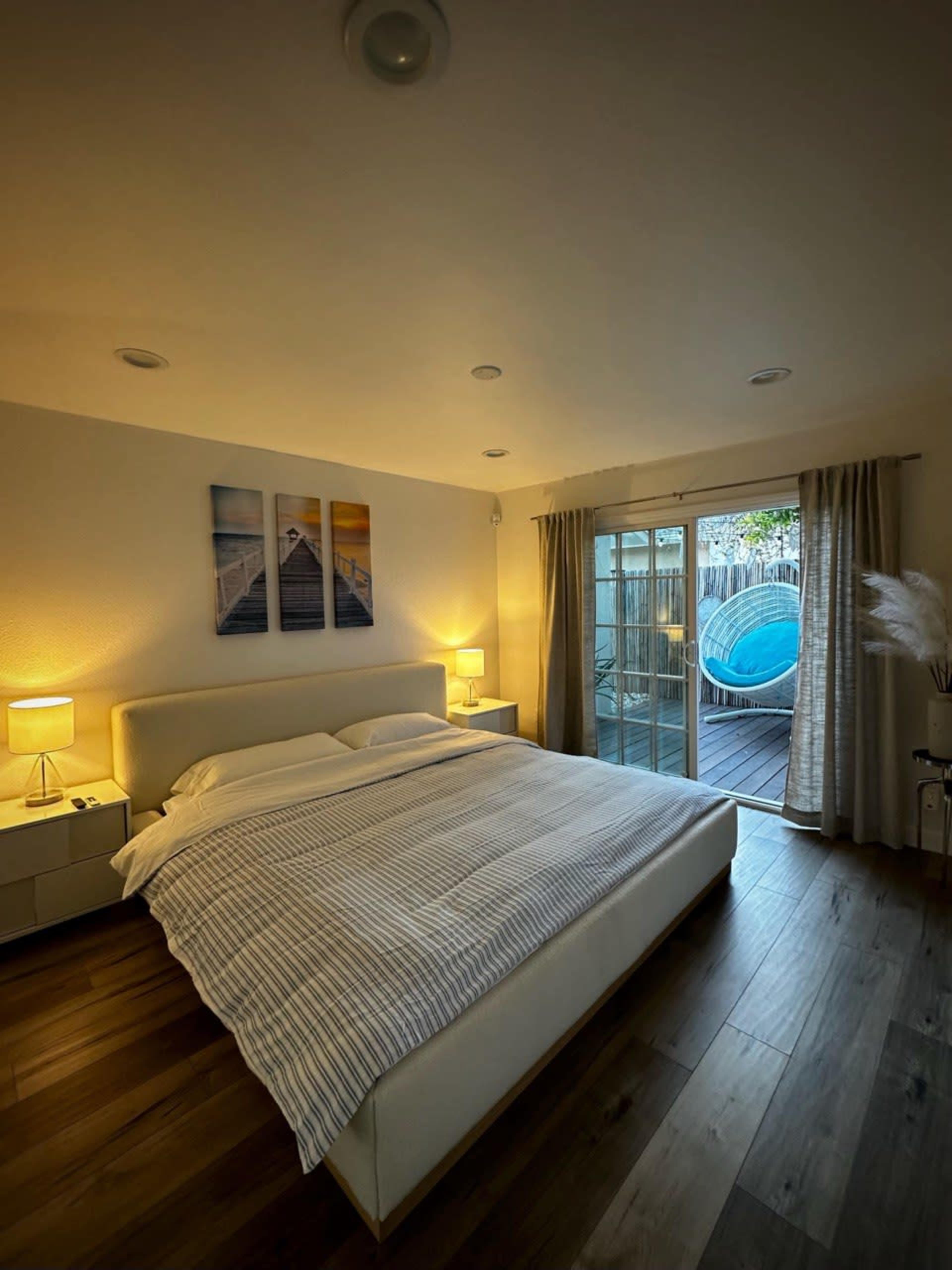 A modern bedroom features a large bed, two lamps, and sliding glass doors leading to an outdoor area with a pool.