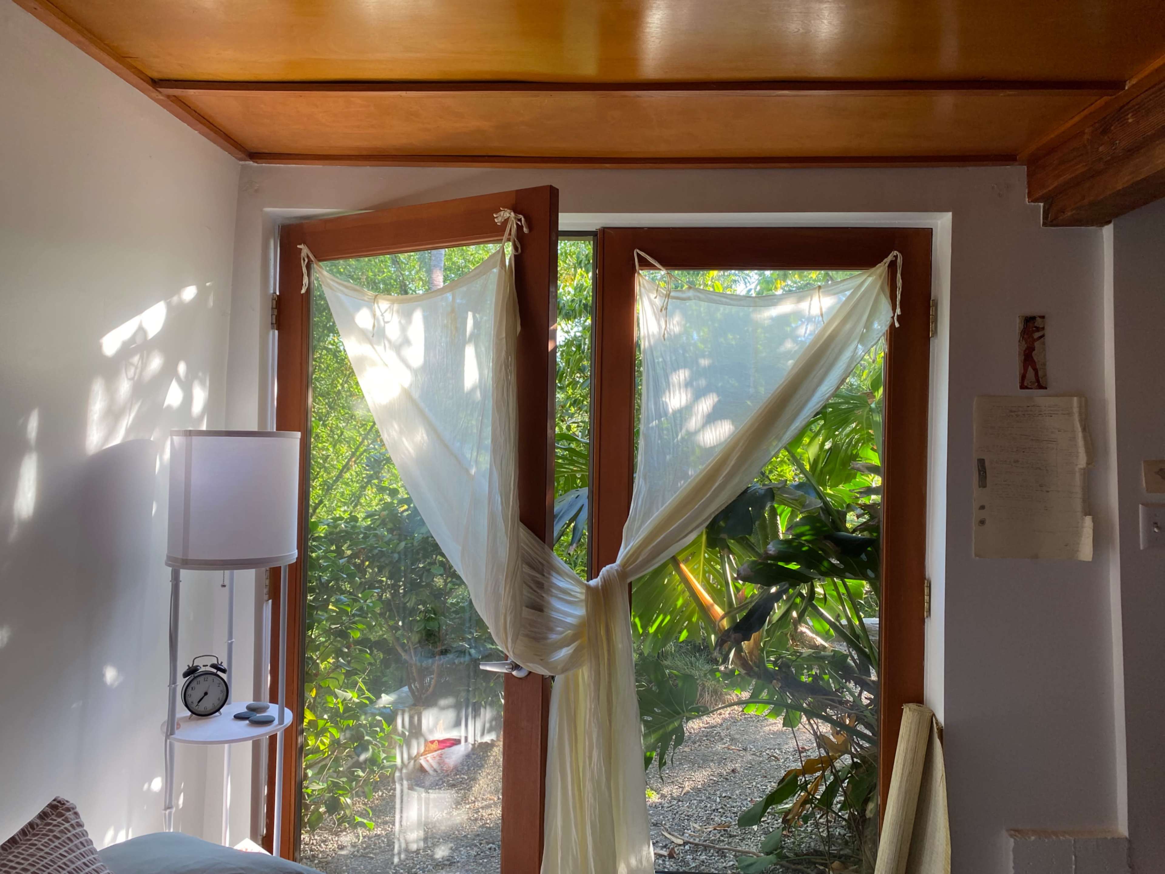 A bright, open room features large wooden windows draped with sheer fabric, overlooking a lush green outdoor area.