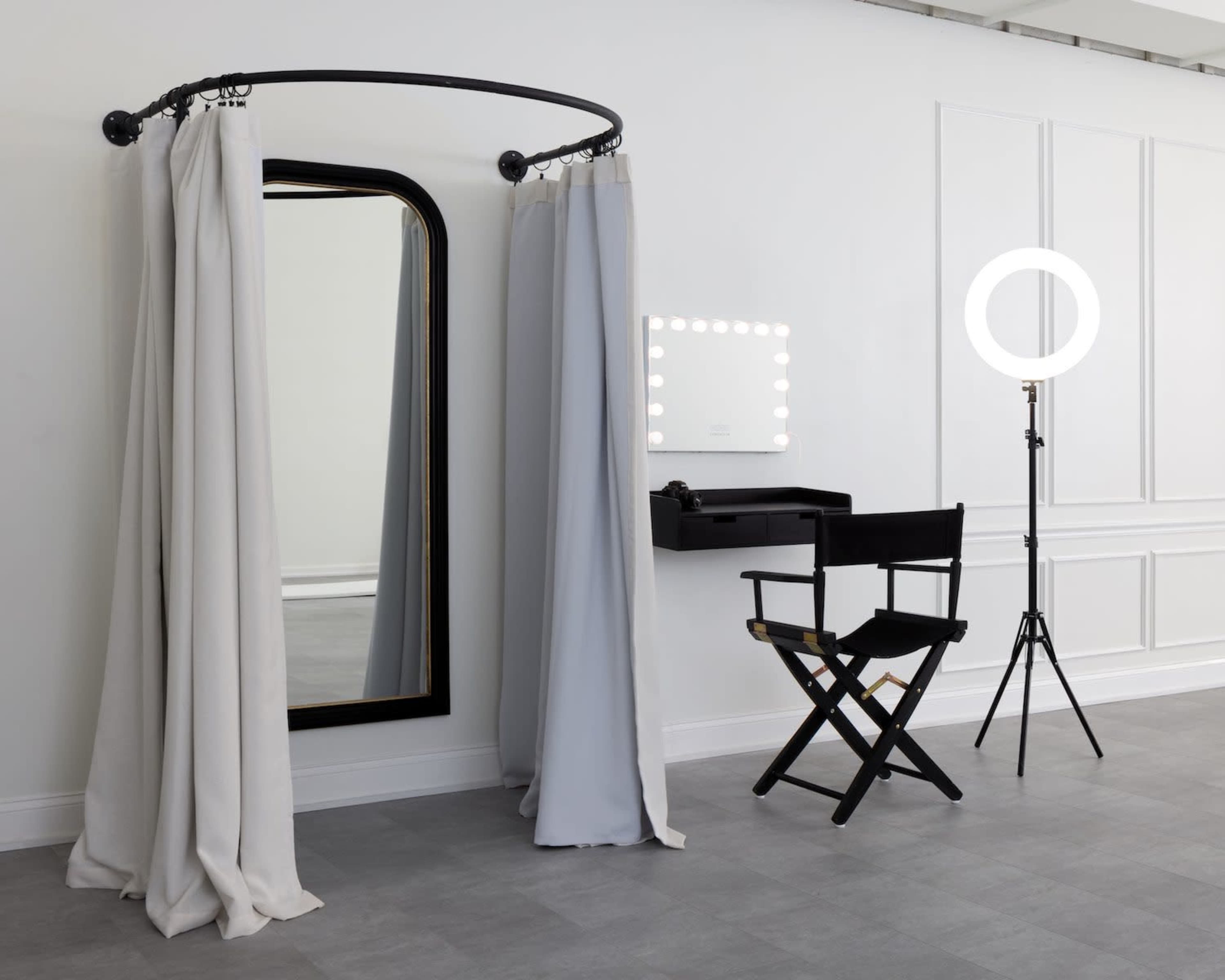 A grooming area featuring a large mirror with lights, a black director's chair, a standing ring light, and draped curtains in a minimalist space.