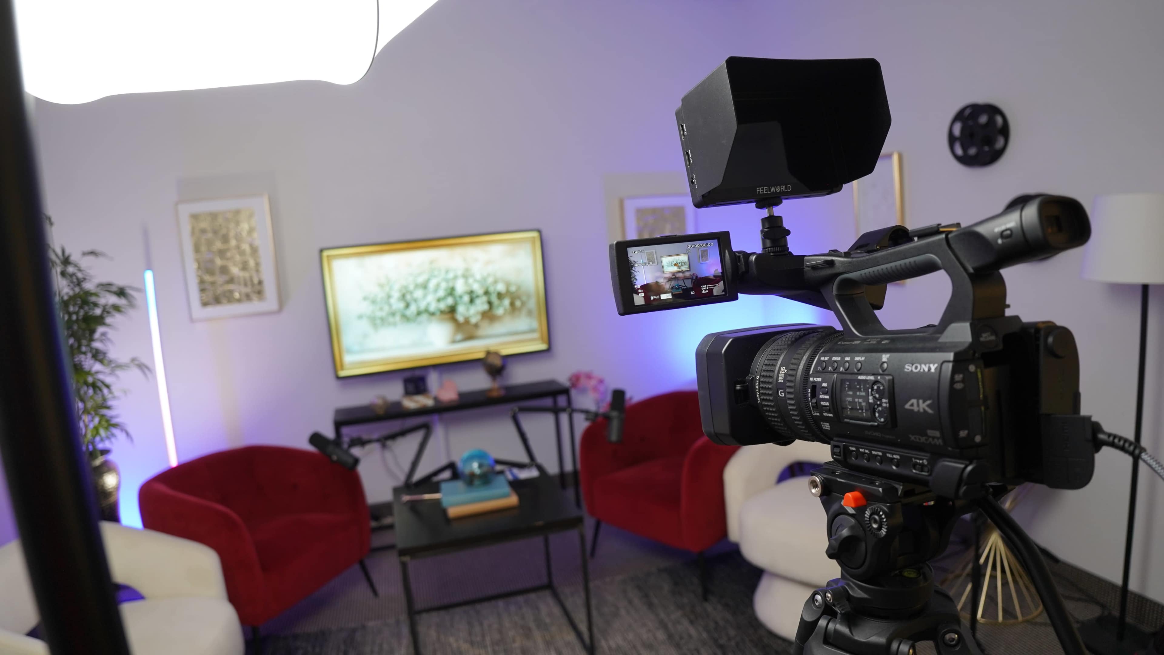 A 4K camera is positioned on a tripod, pointed towards a decorated seating area with red chairs, a coffee table, and a television displaying artwork.