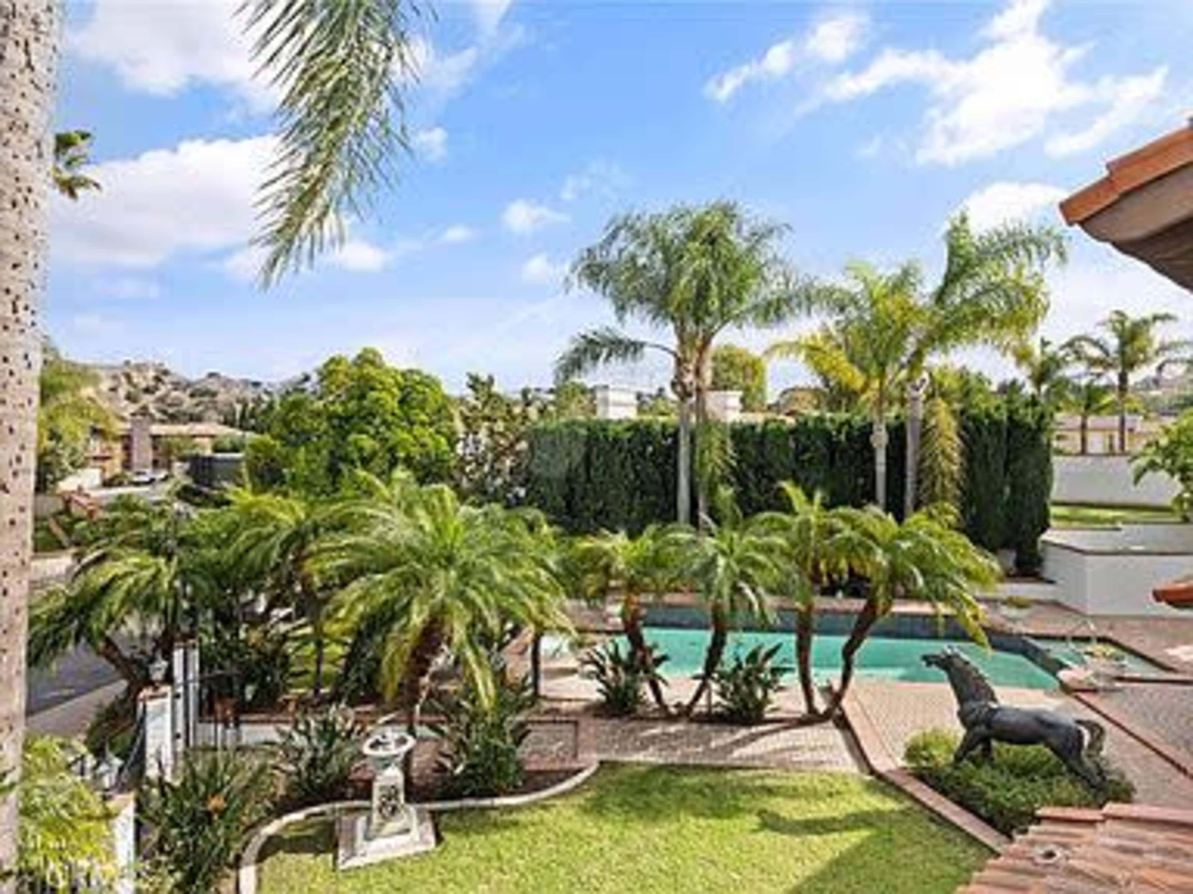 The image shows a landscaped backyard featuring a pool surrounded by palm trees and a lion statue.
