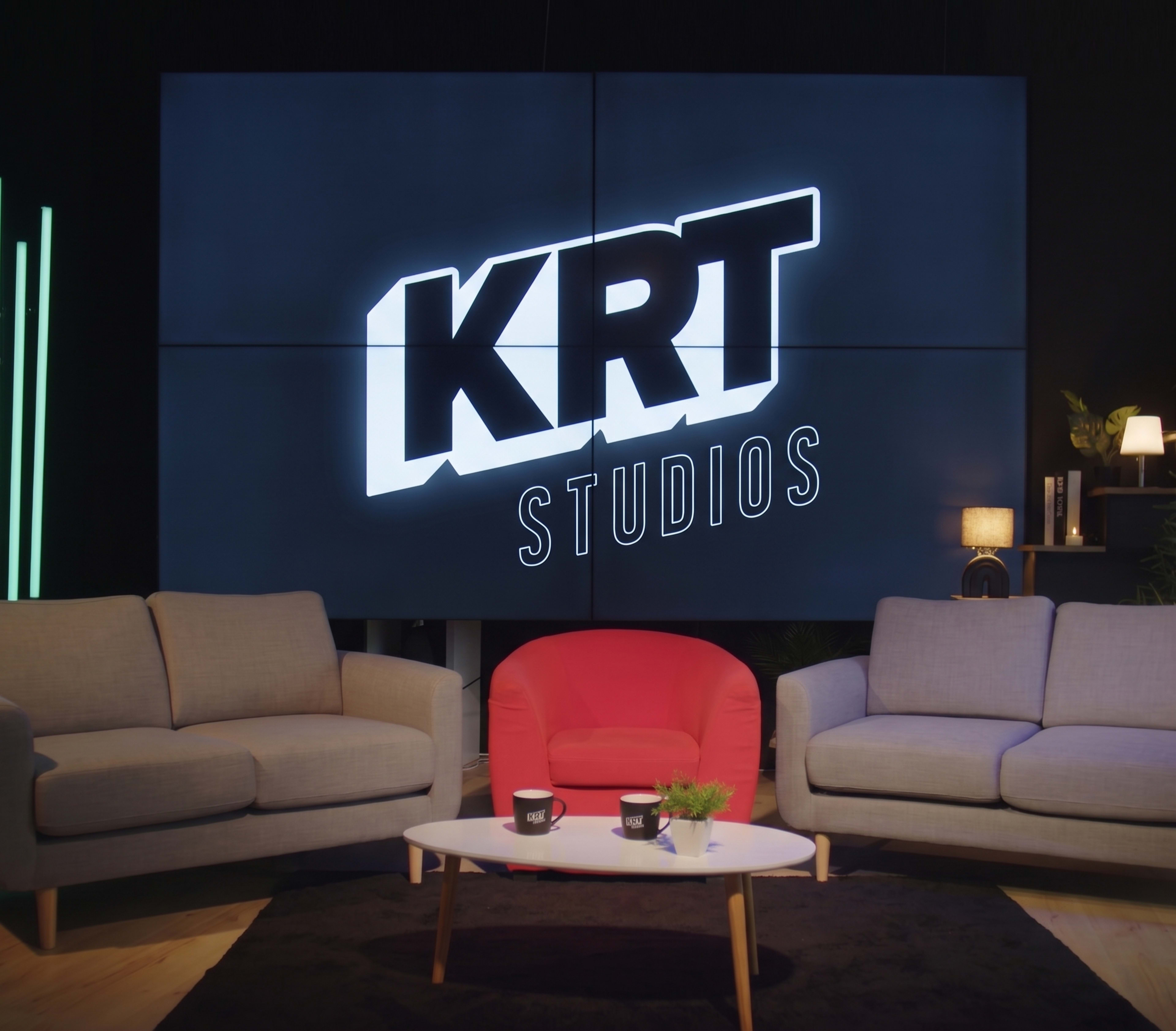The image shows a studio set with two sofas, a coffee table, and a large screen displaying the logo "KRT STUDIOS" in the background.