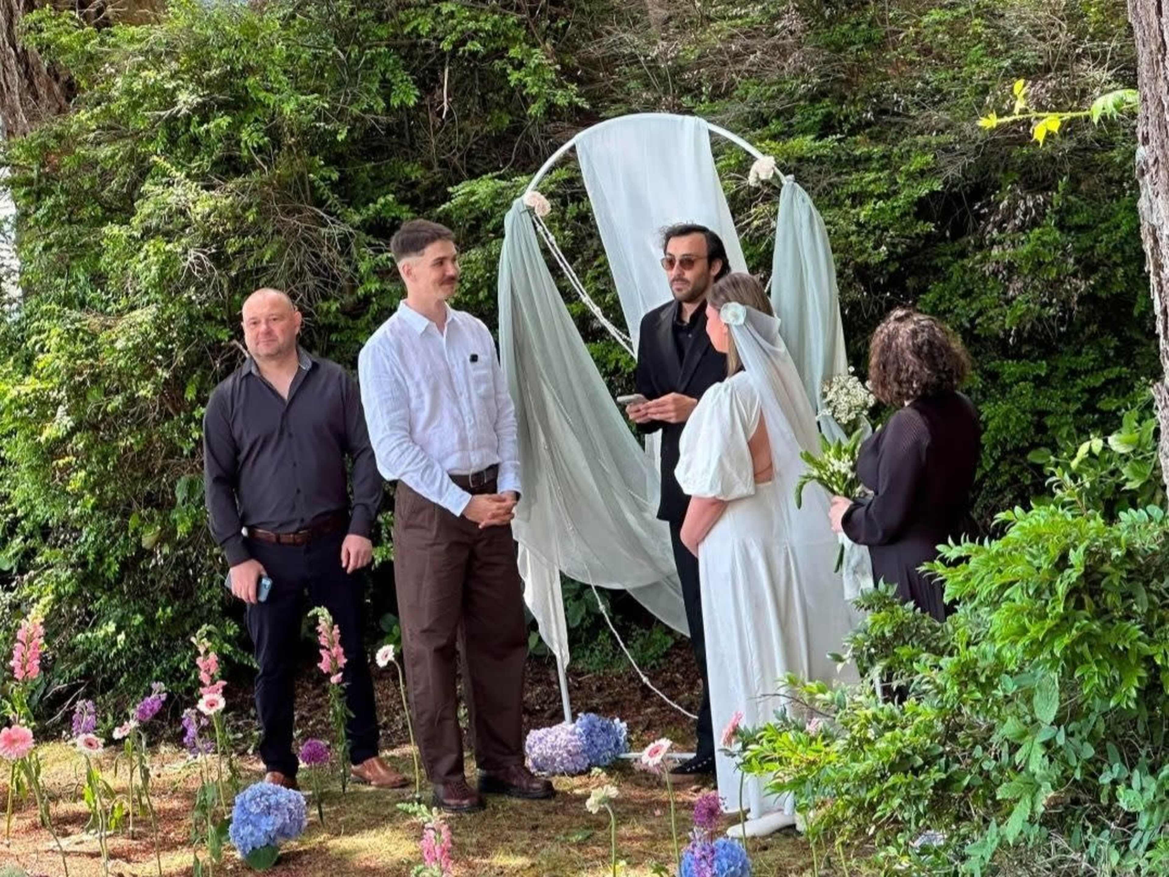 A wedding ceremony is taking place outdoors with a couple exchanging vows in front of a floral arch, while four guests observe.