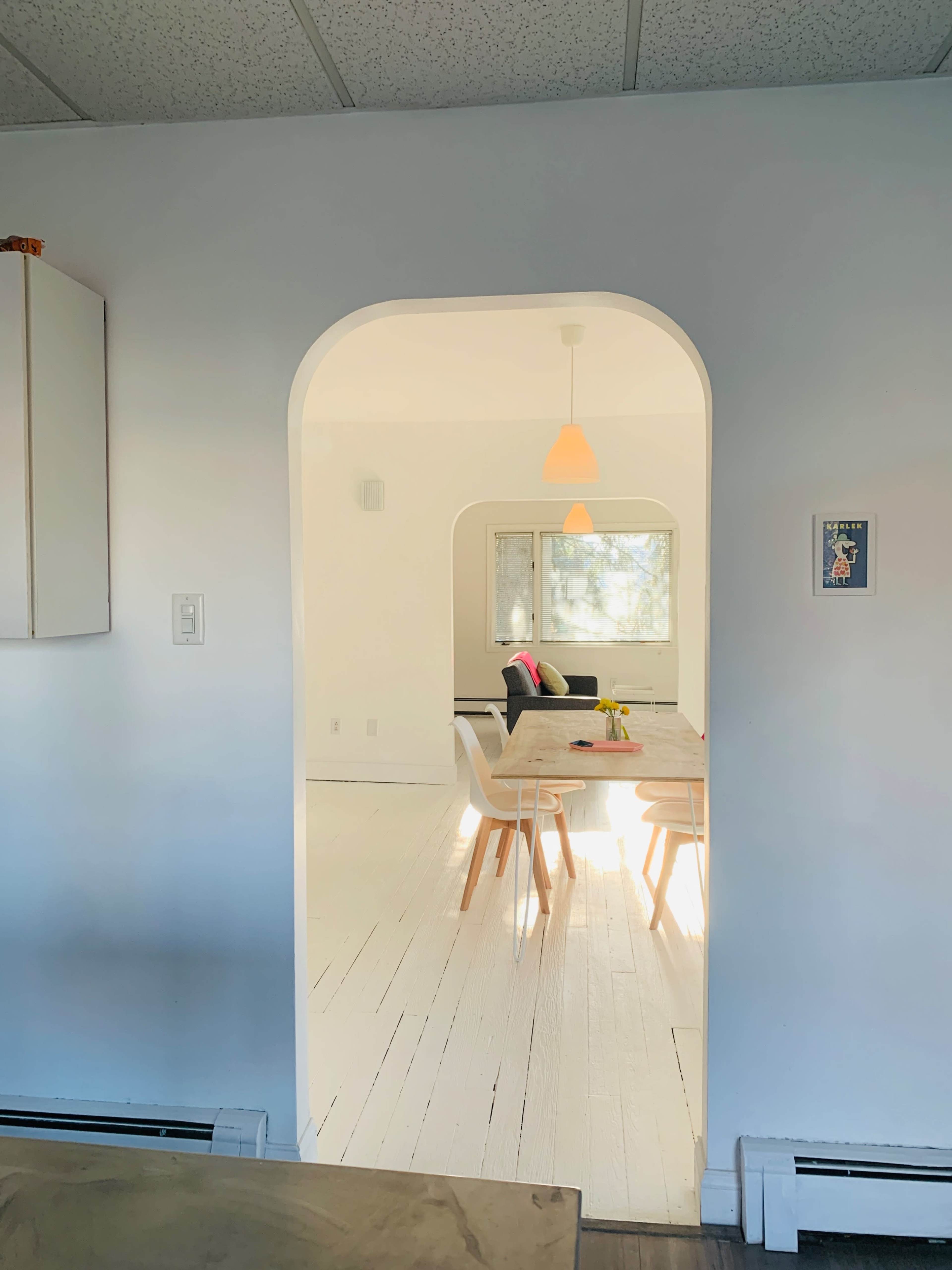 A simple archway leads from a kitchen area into a bright, open living space furnished with a dining table and chairs.