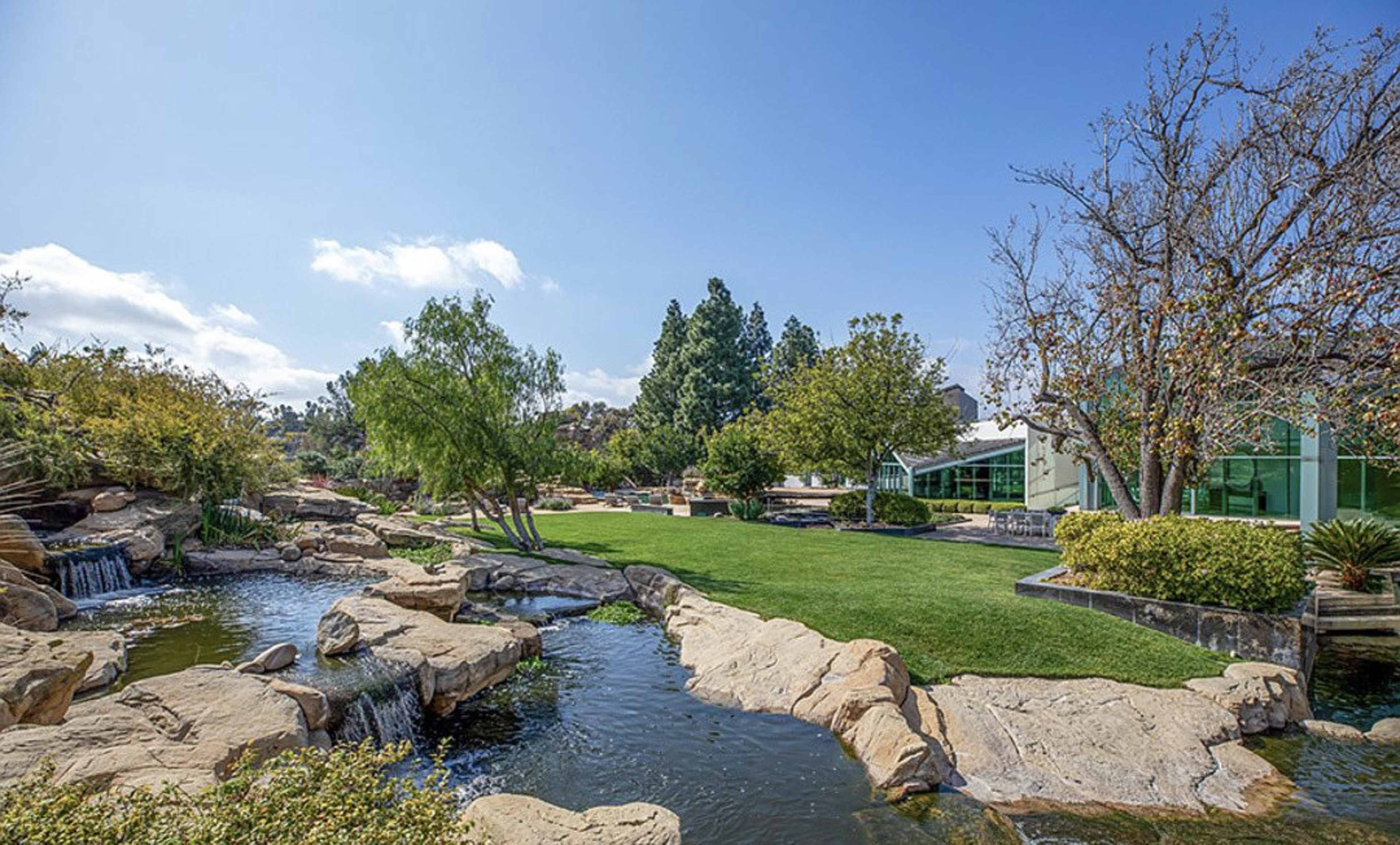The scene features a landscaped area with a flowing stream, rocks, waterfalls, and a green lawn, accompanied by trees and a modern building in the background.