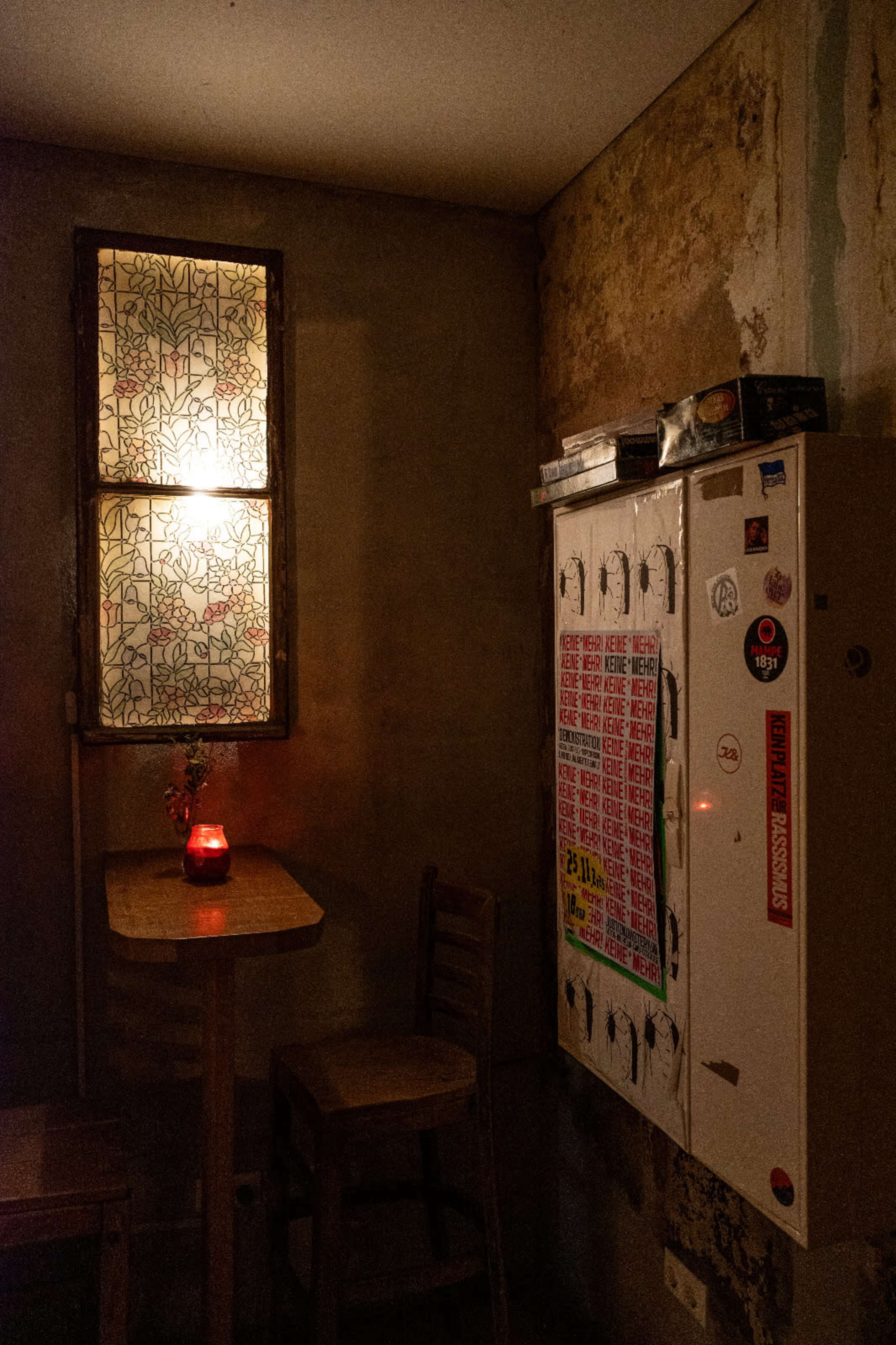 A small, dimly lit corner of a room features a wooden table and chair next to a decorative window and a sticker-covered cabinet.