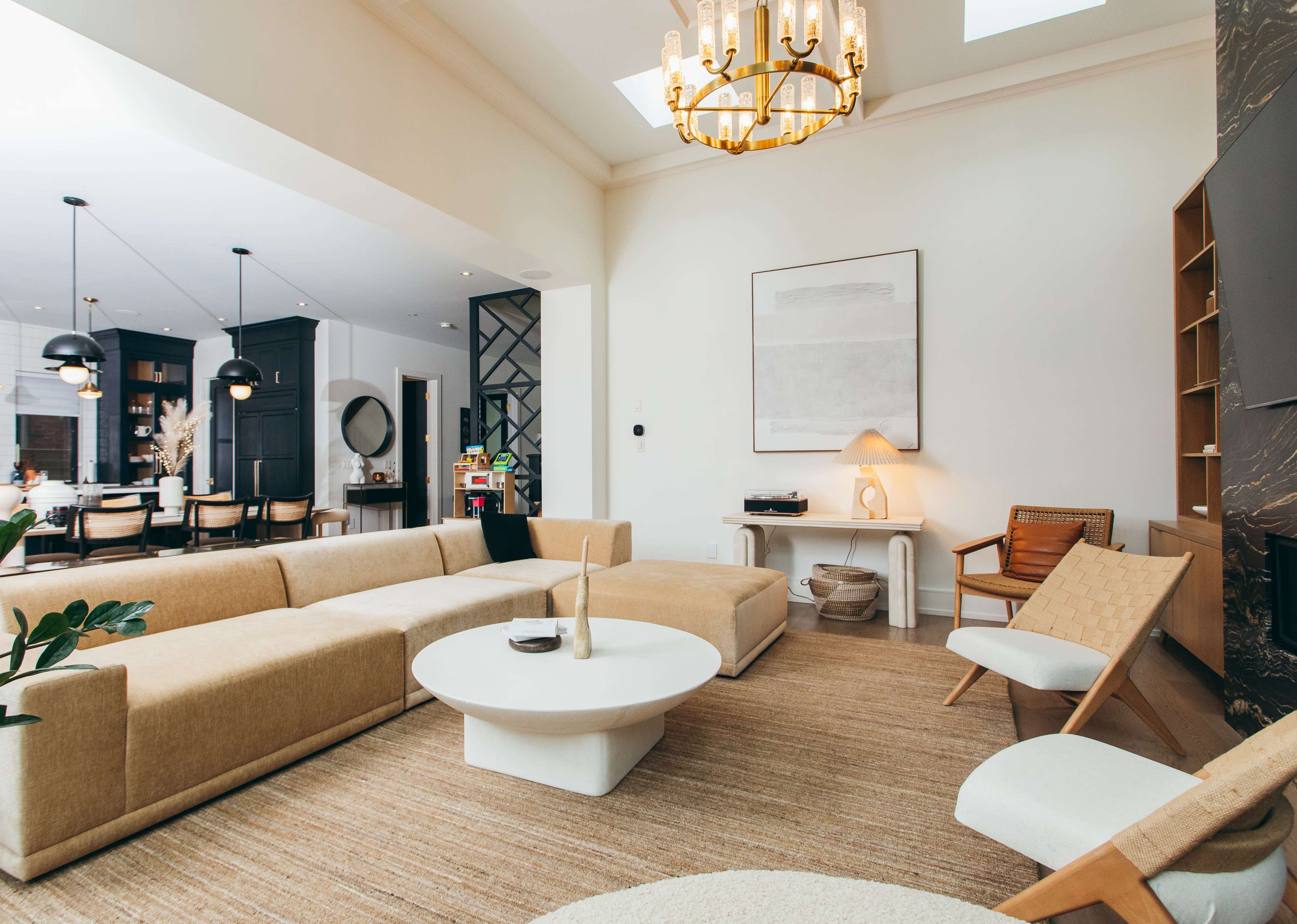The image shows a modern living room with a large beige sectional sofa, a round white coffee table, and light-colored furniture, complemented by a chandelier and a minimalistic artwork on the wall.