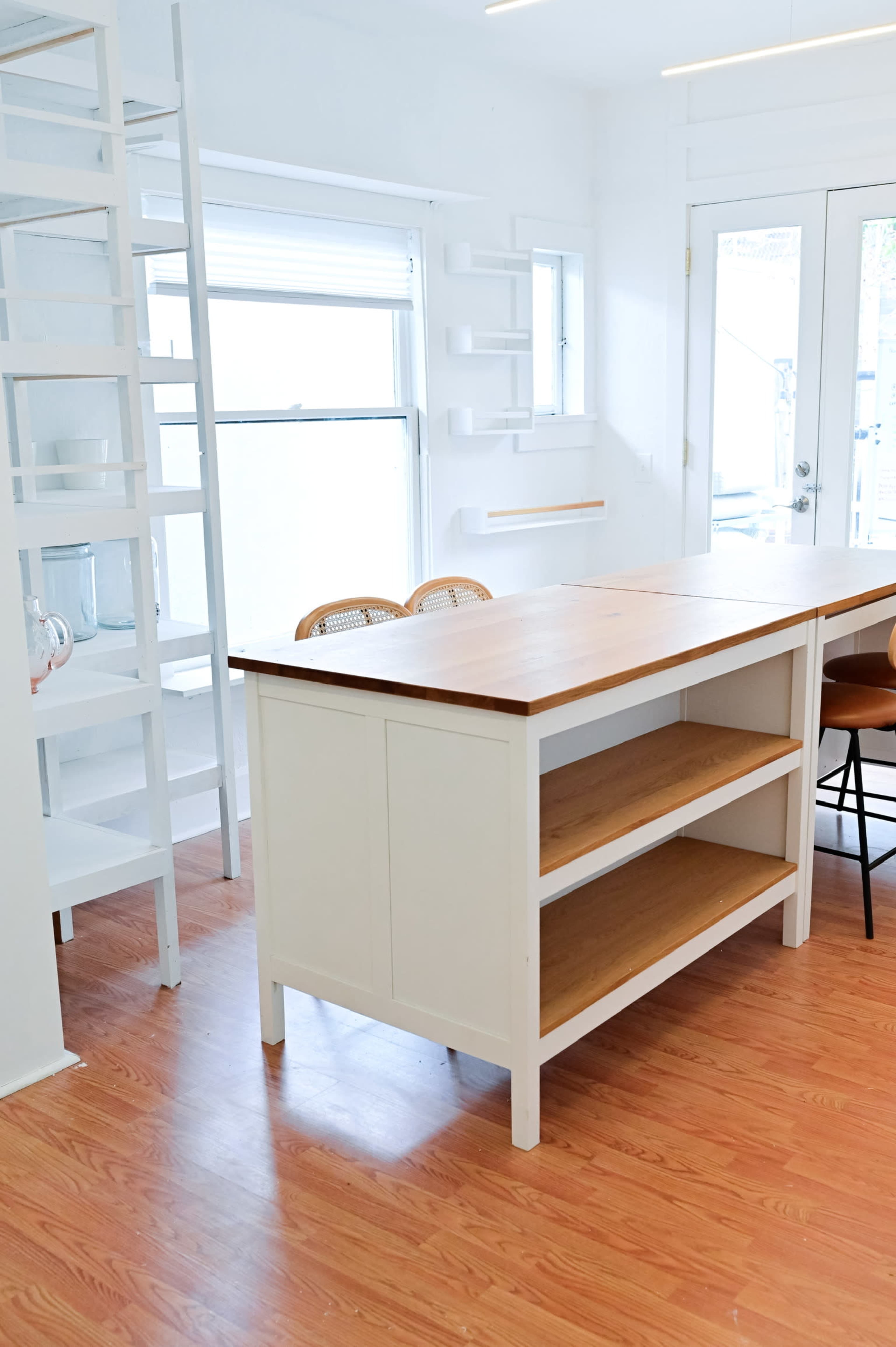 A bright kitchen features a white island with wooden shelves and a tall ladder-style shelf in the background.
