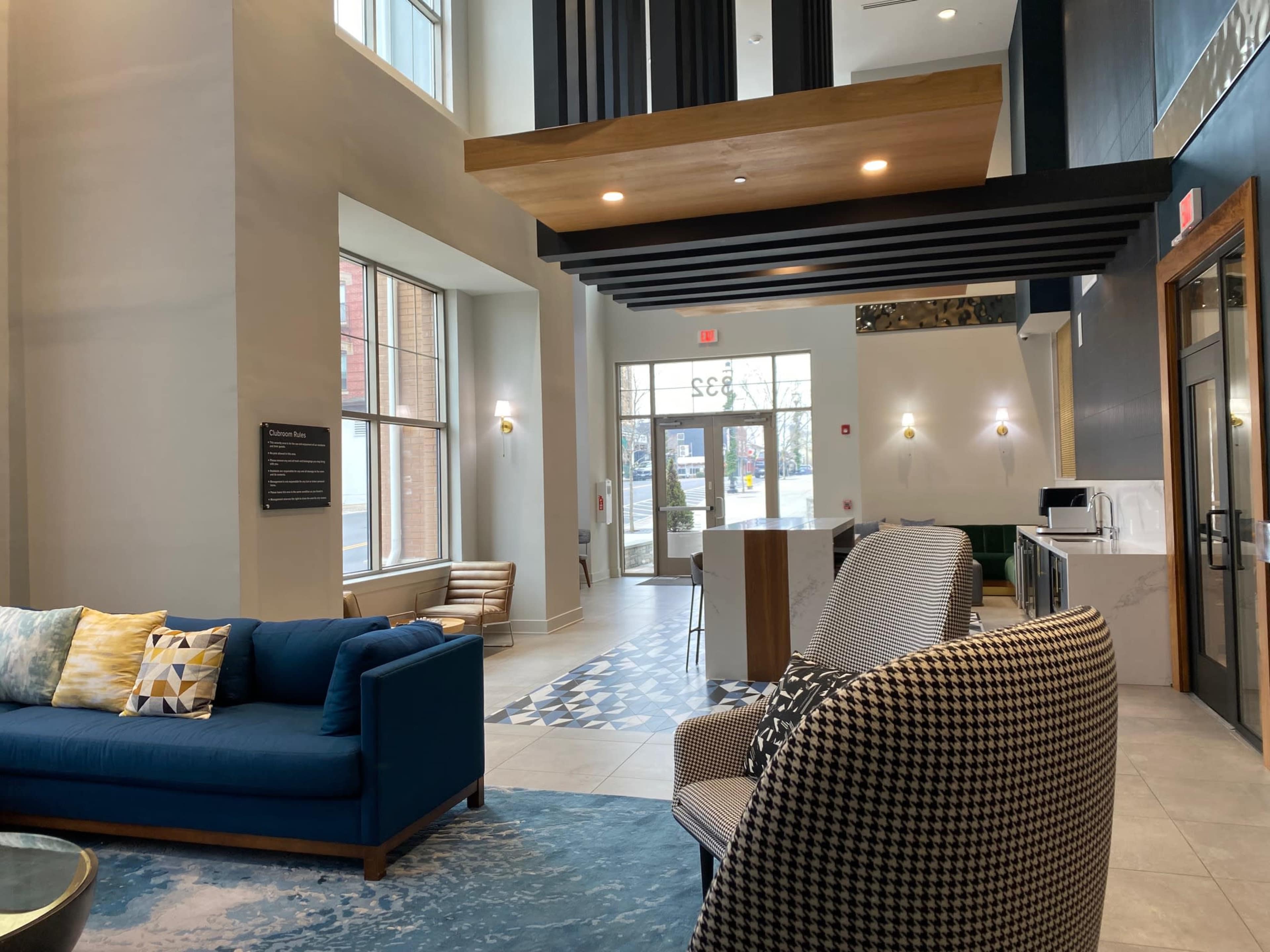 The image shows a modern lobby with a blue couch, patterned chairs, and large windows allowing natural light to enter the space.