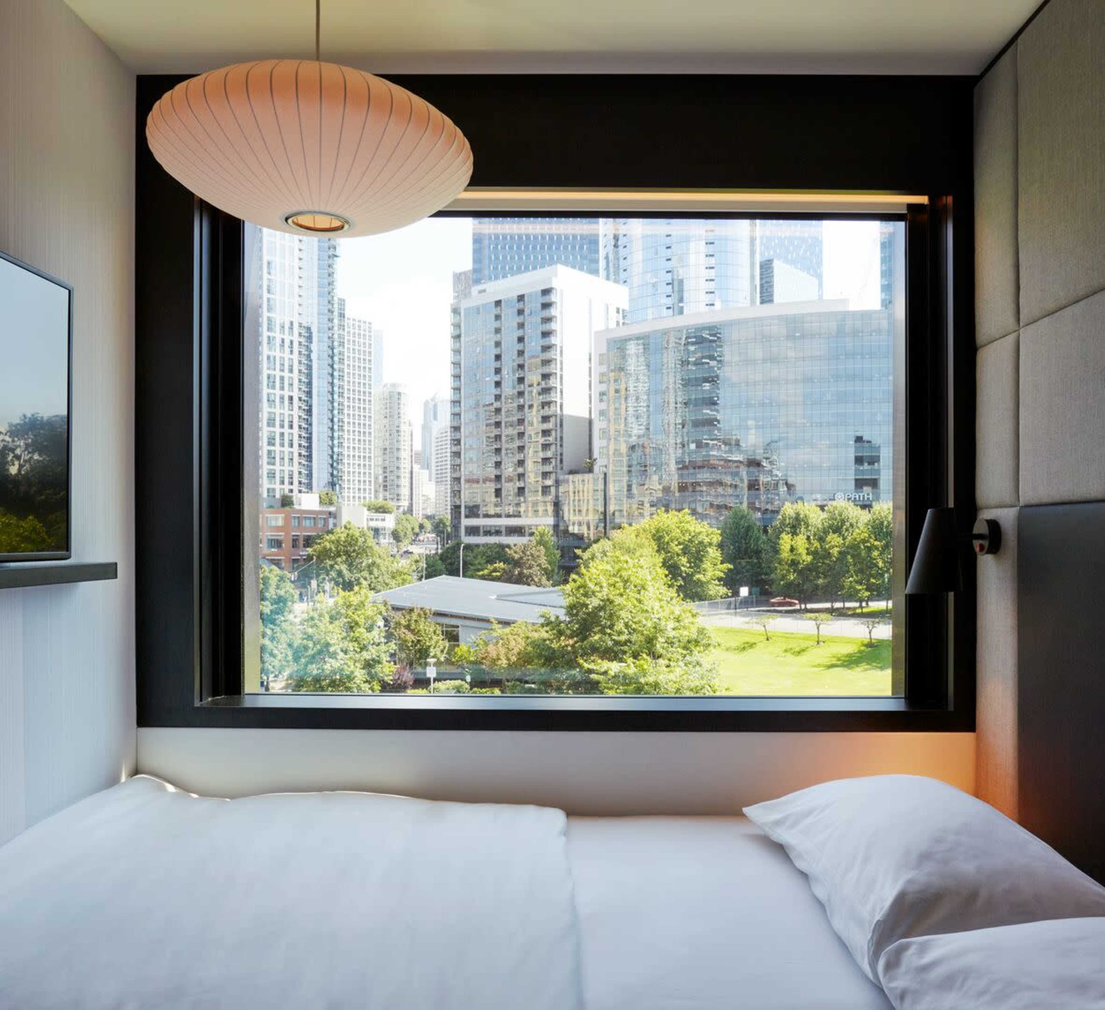 A hotel room features a large window overlooking a cityscape with modern buildings and a park.
