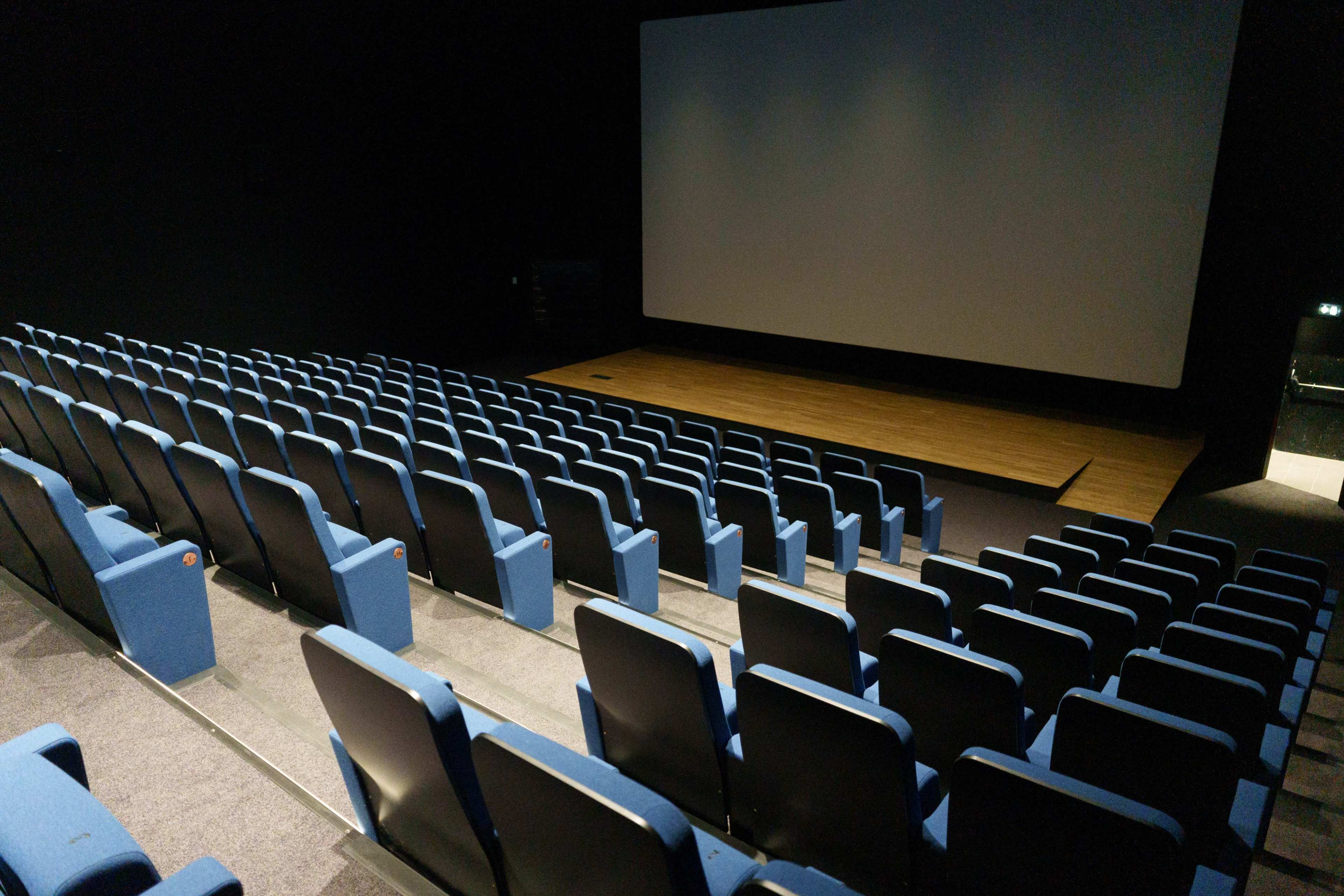 An empty movie theater features rows of blue seats facing a large, blank screen.