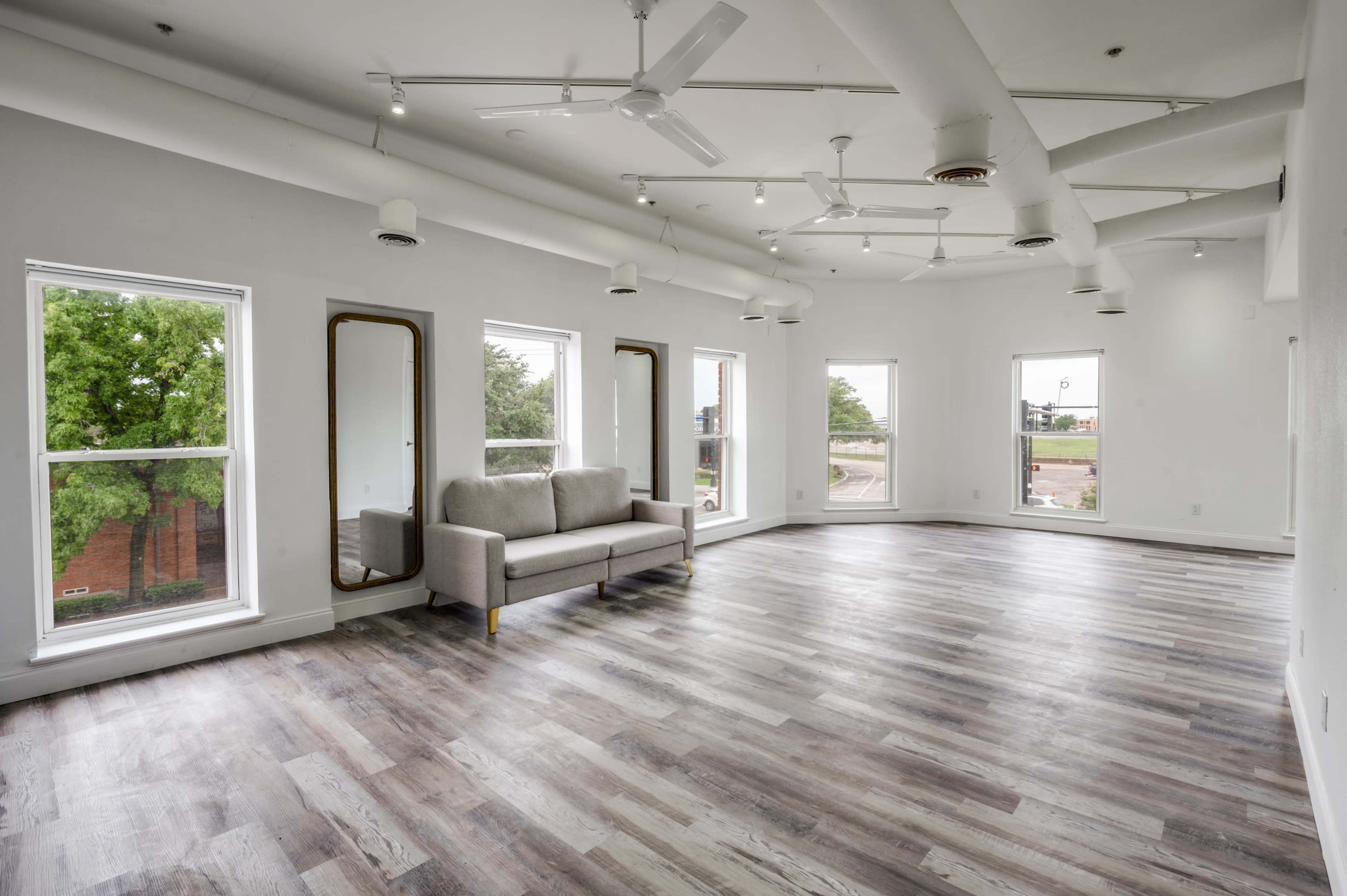 A spacious, well-lit room with hardwood flooring, two large windows, a gray couch, and mirrors on the walls.