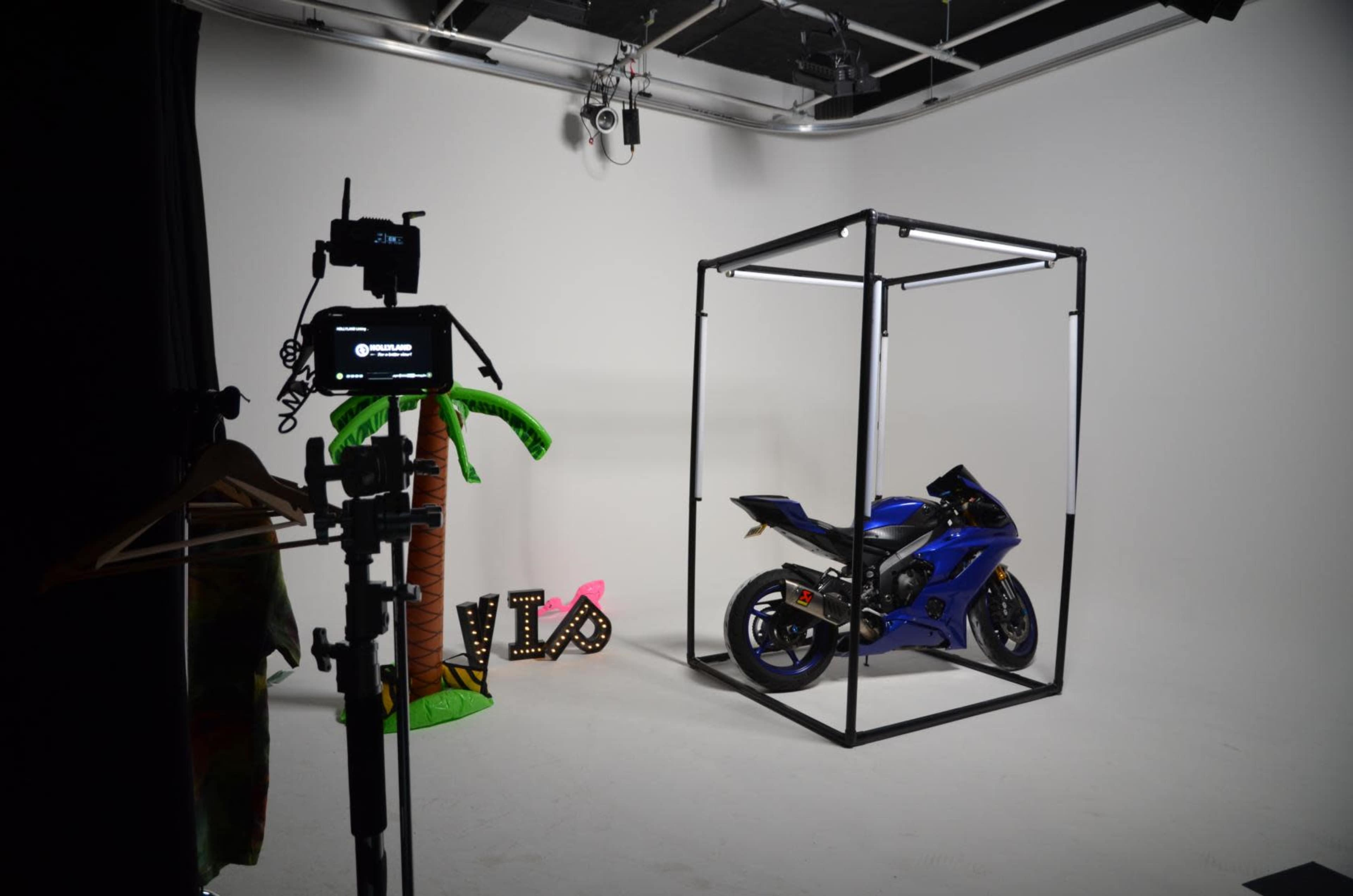 A blue motorcycle is displayed inside a metal frame on a plain background, with a palm tree decoration and a "VIP" sign nearby.