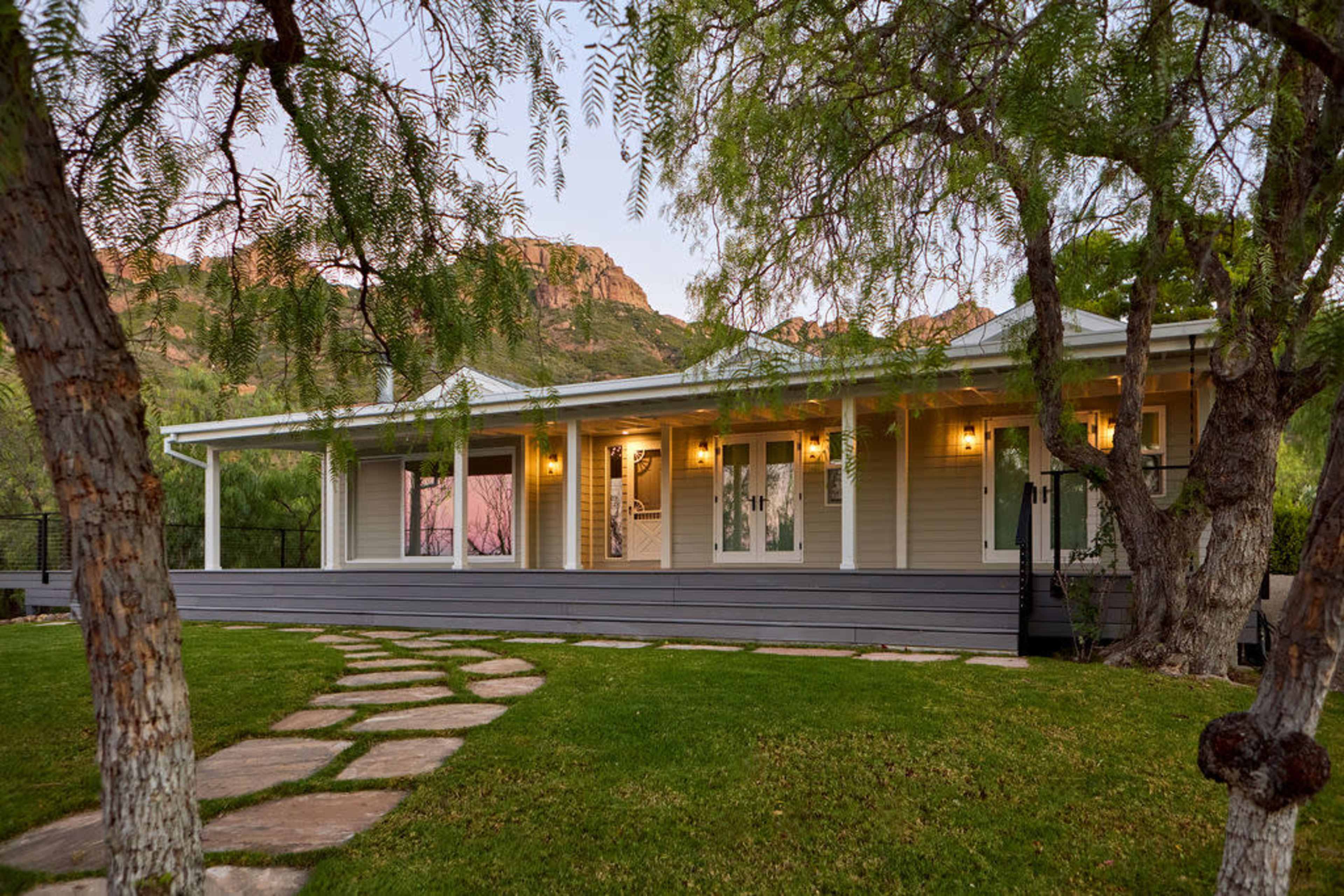 A single-story house with a front porch and large windows is set against a backdrop of mountains, surrounded by green grass and trees.