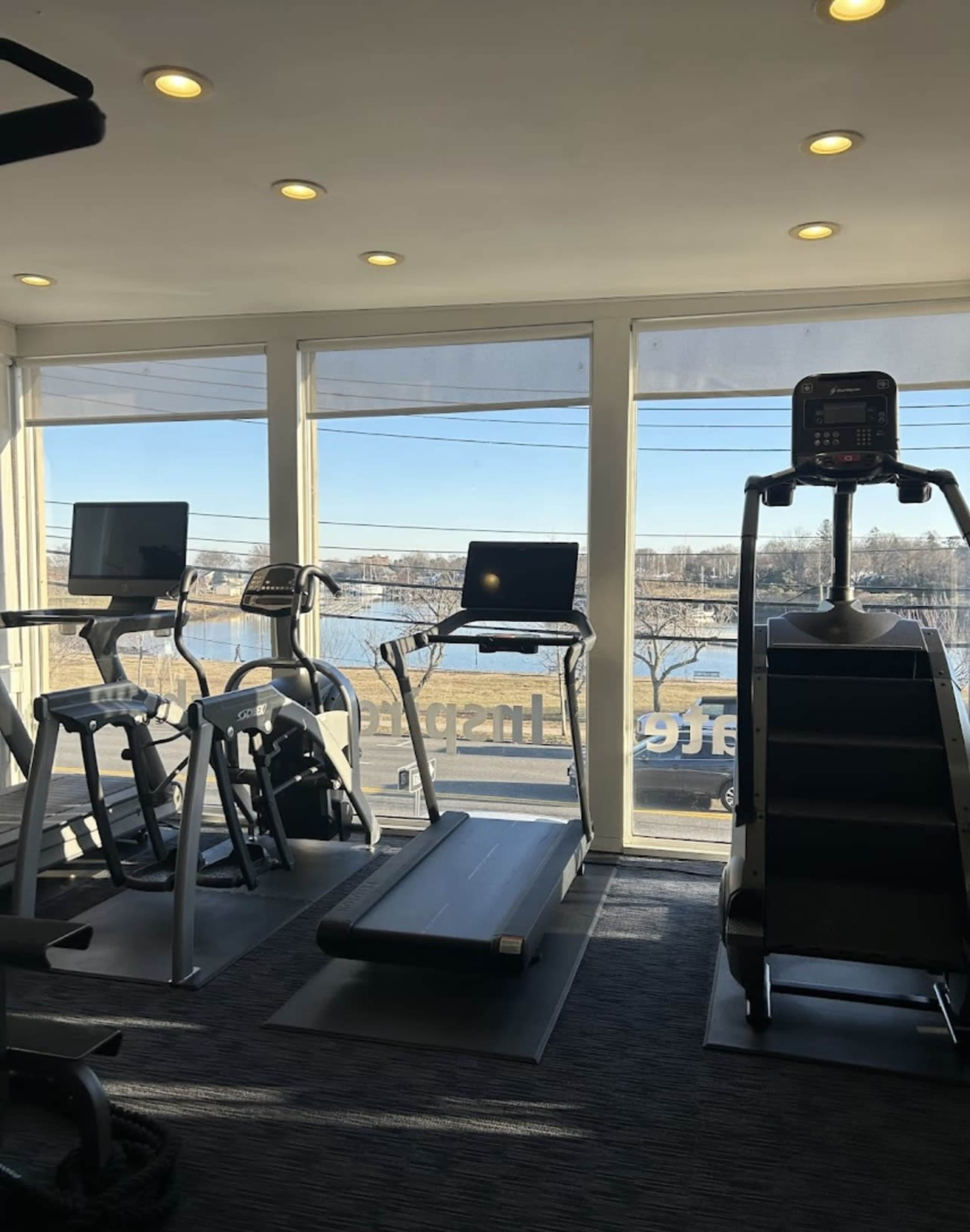 The gym features several workout machines, including treadmills and stair climbers, with large windows showcasing a view of the river outside.