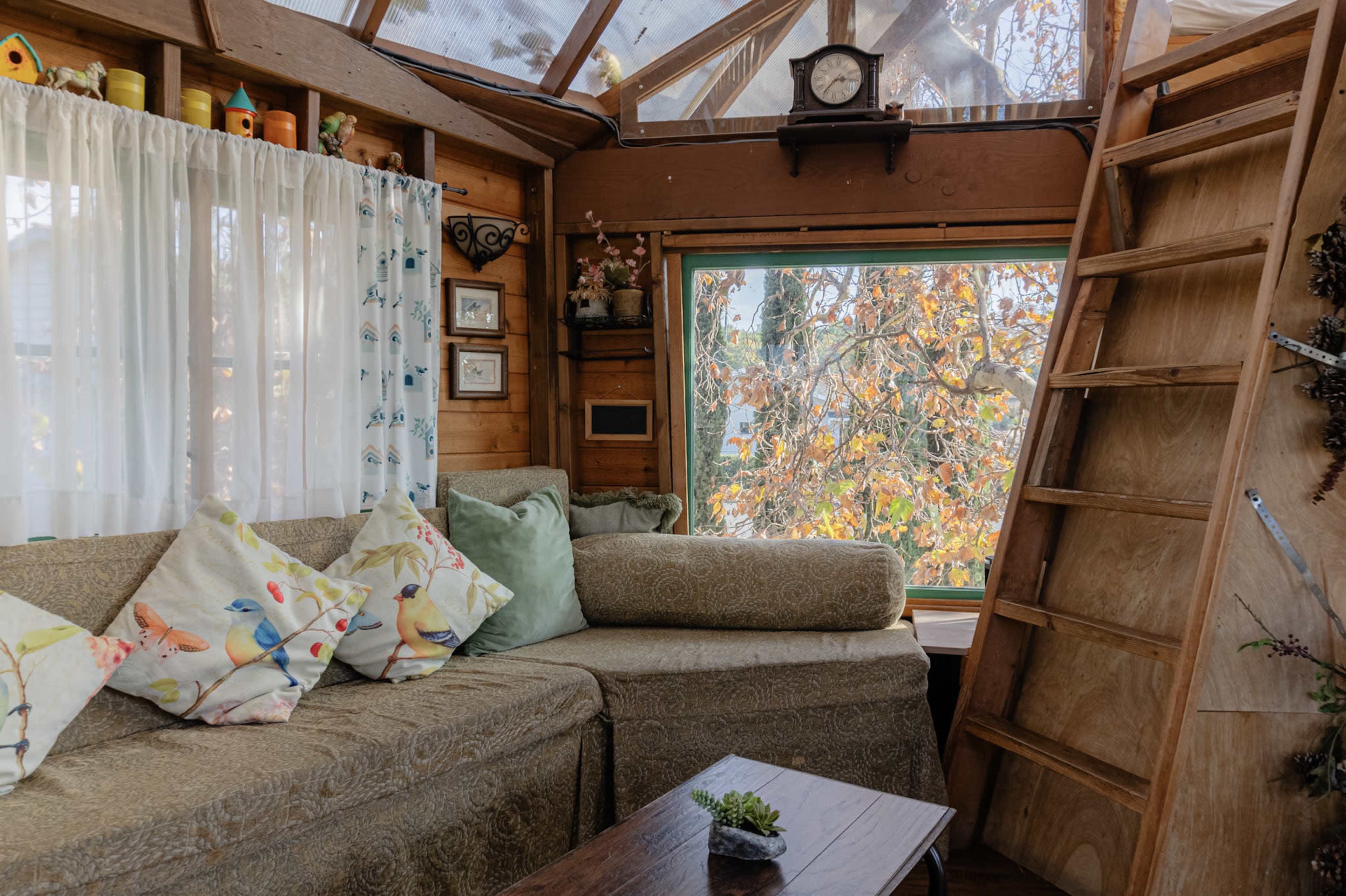 A cozy interior features a sectional couch with patterned pillows, set against a large window overlooking autumn foliage, with a wooden ladder leading to an upper level.
