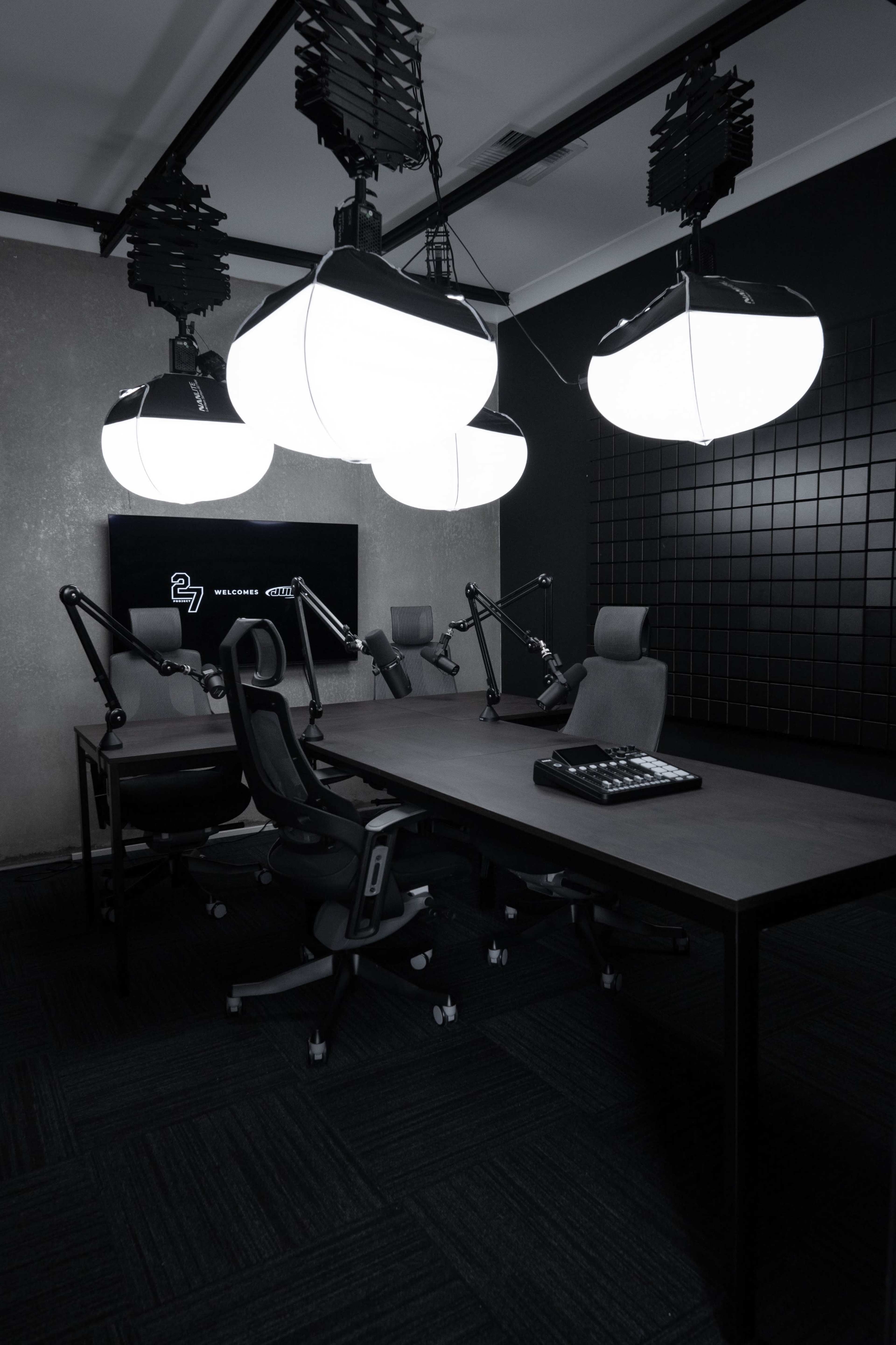 The image shows a modern recording studio equipped with a large table, adjustable chairs, and overhead lighting.