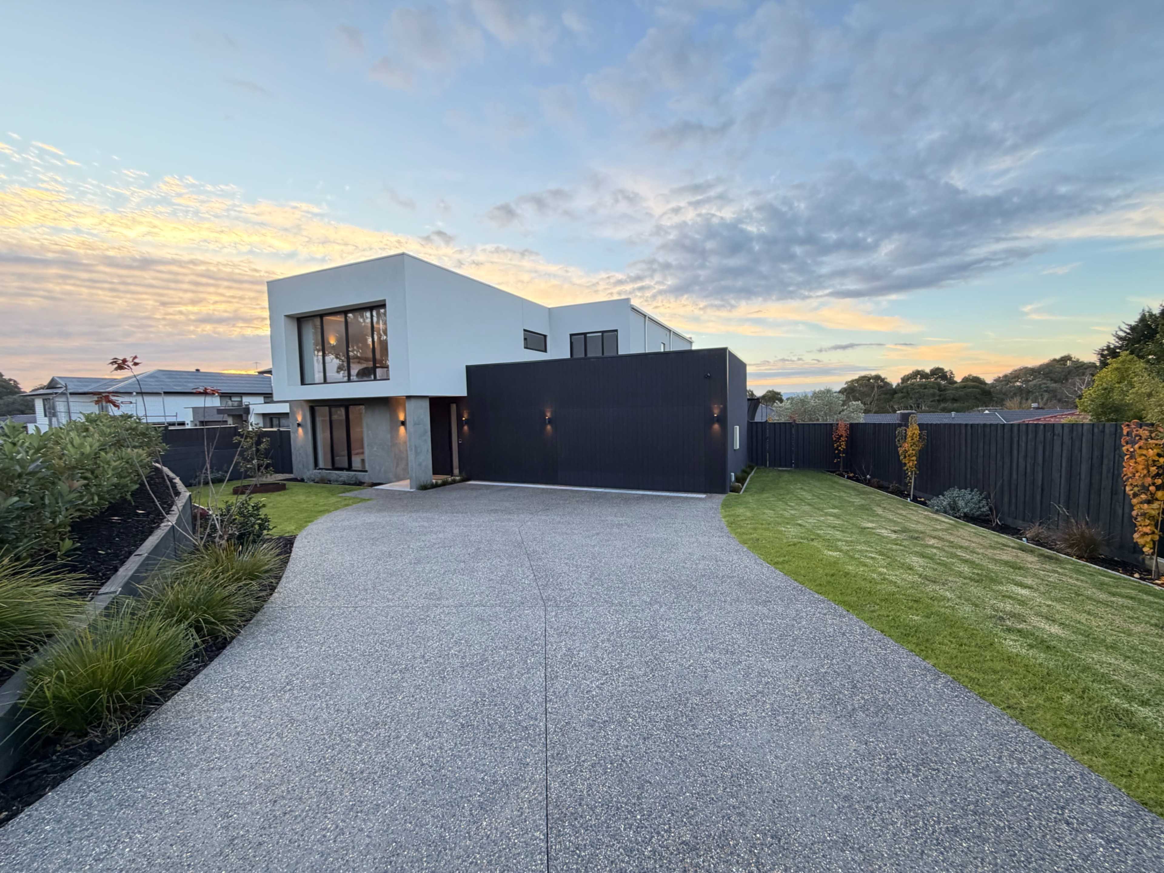 The image shows a modern house with a predominately white exterior and a black accent wall, situated on a gravel driveway and surrounded by landscaped greenery.