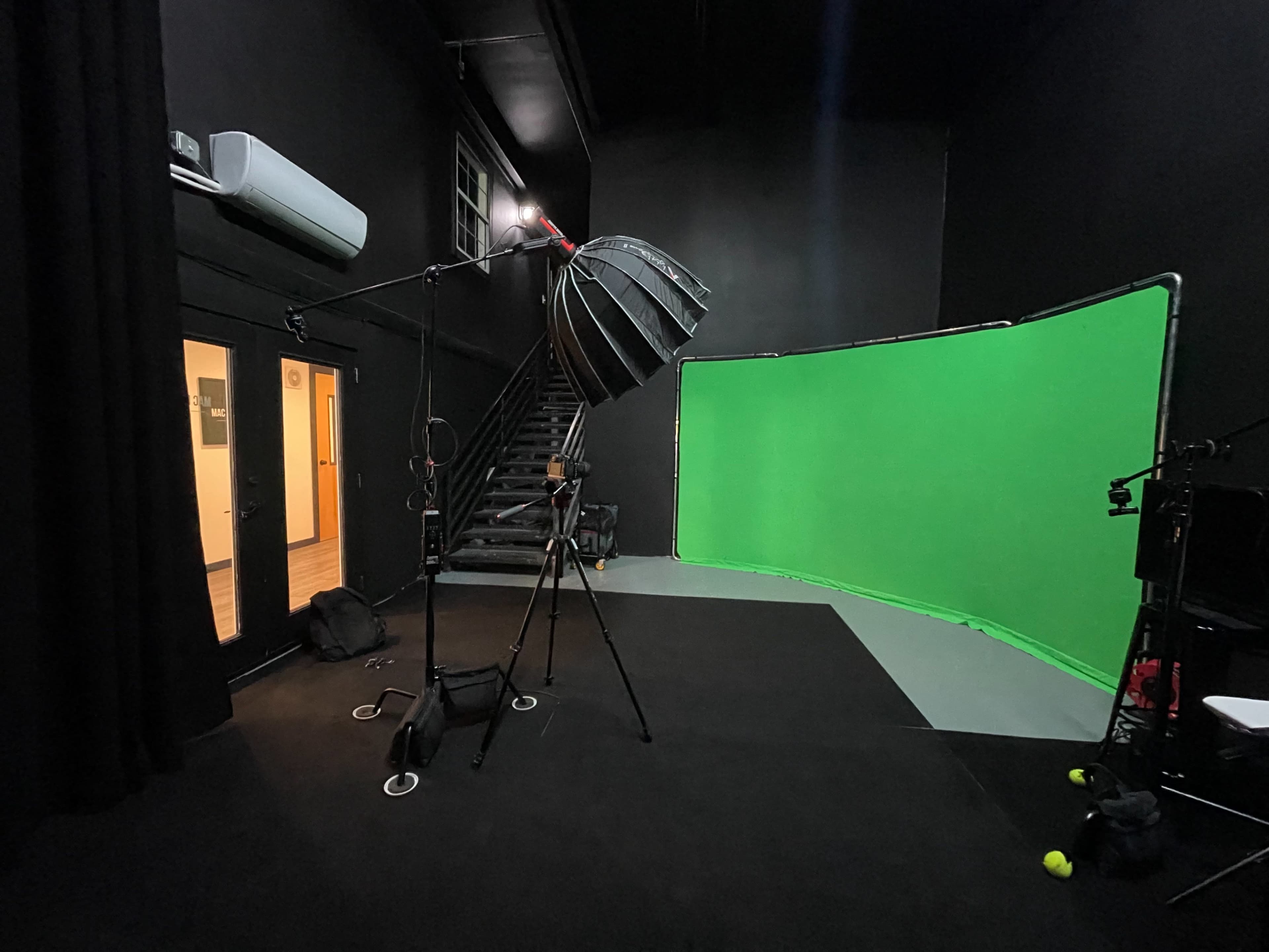 A photography studio features a green screen, a large light mounted on a stand, and stairs leading to a door.
