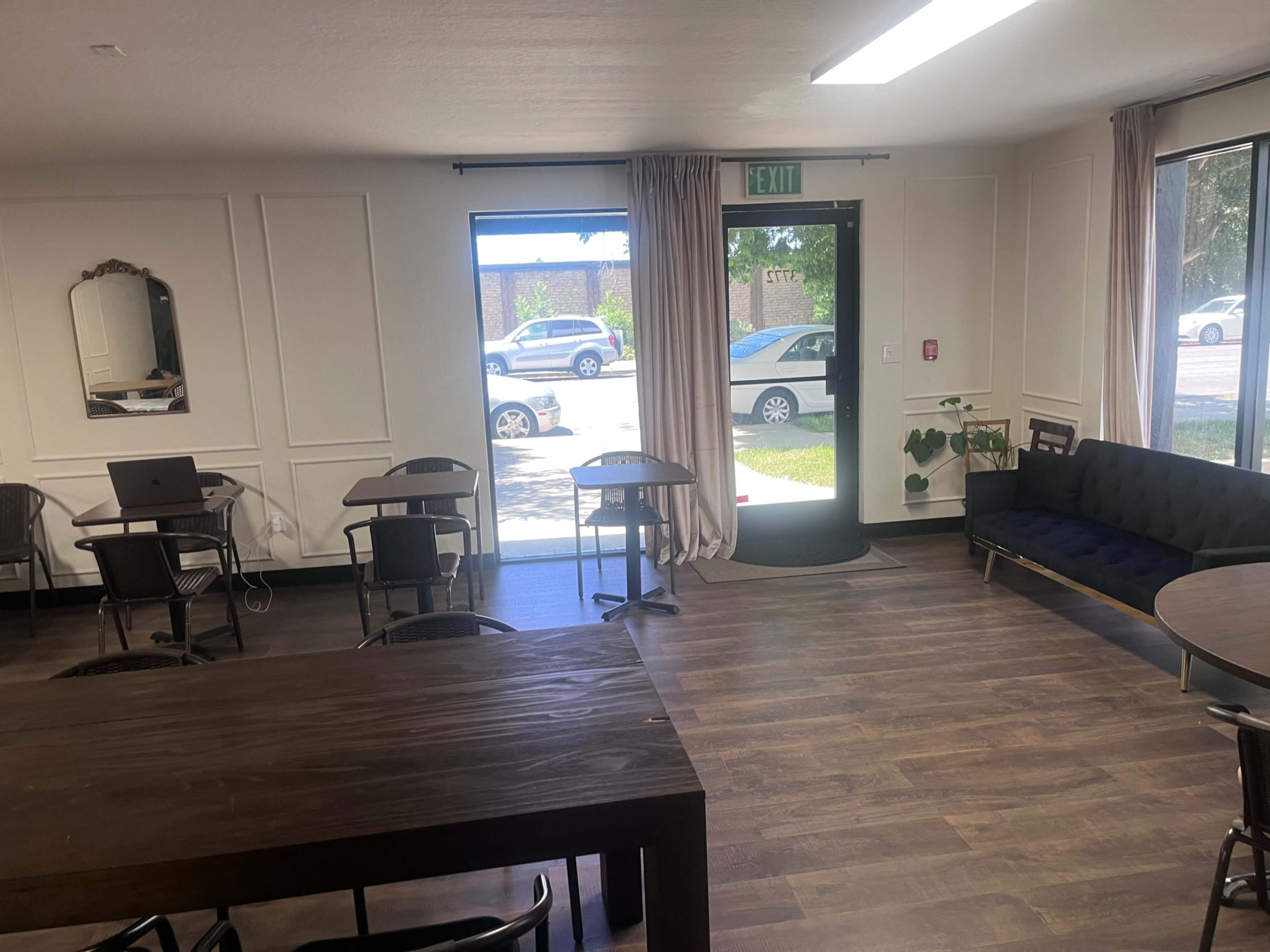 The image shows a spacious room with wooden flooring, featuring several tables and chairs, a couch, and large windows that provide a view of parked cars outside.