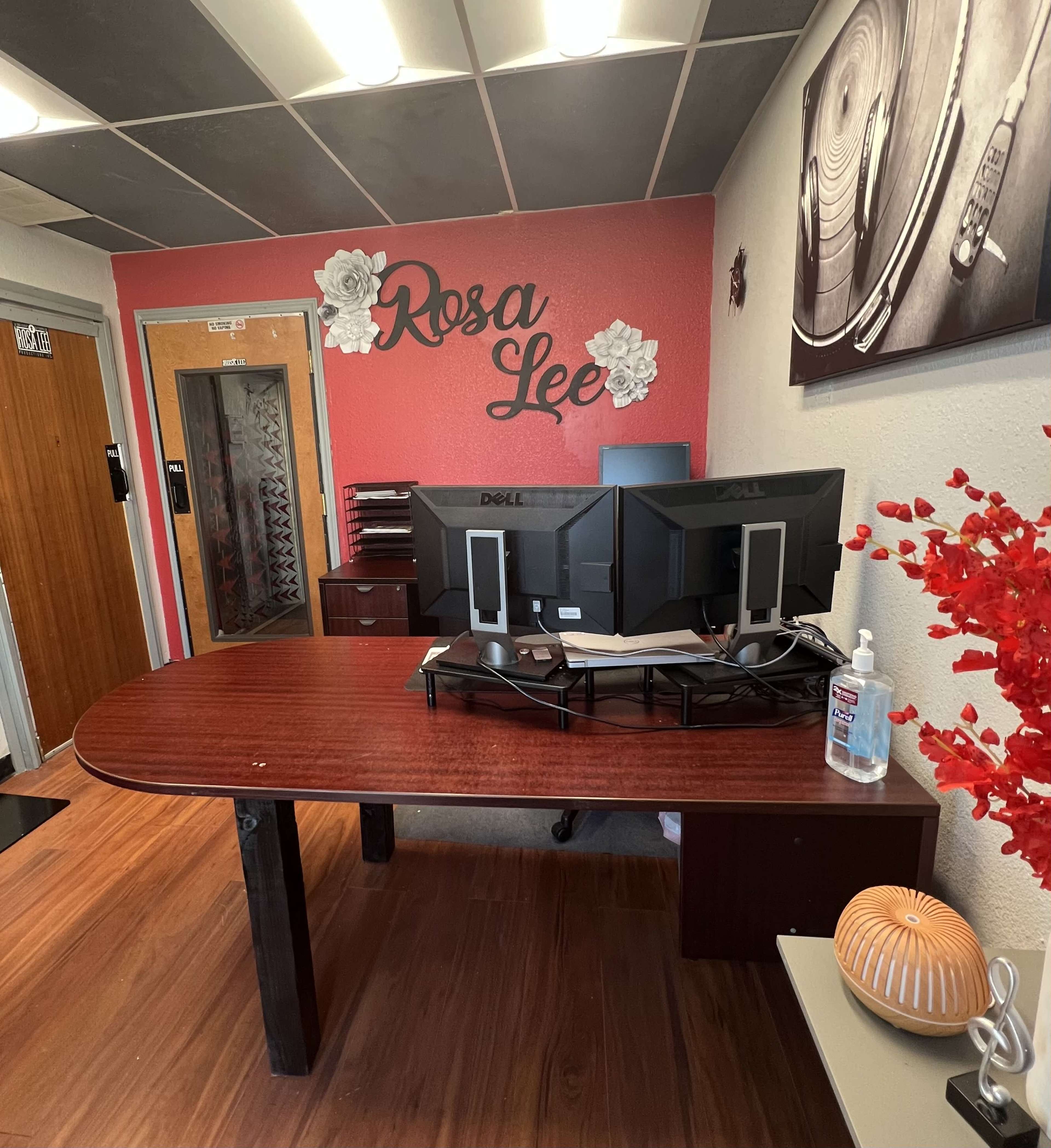 The image shows an office reception area featuring a wooden desk with two Dell monitors, a hand sanitizer dispenser, and decorative elements on the walls.