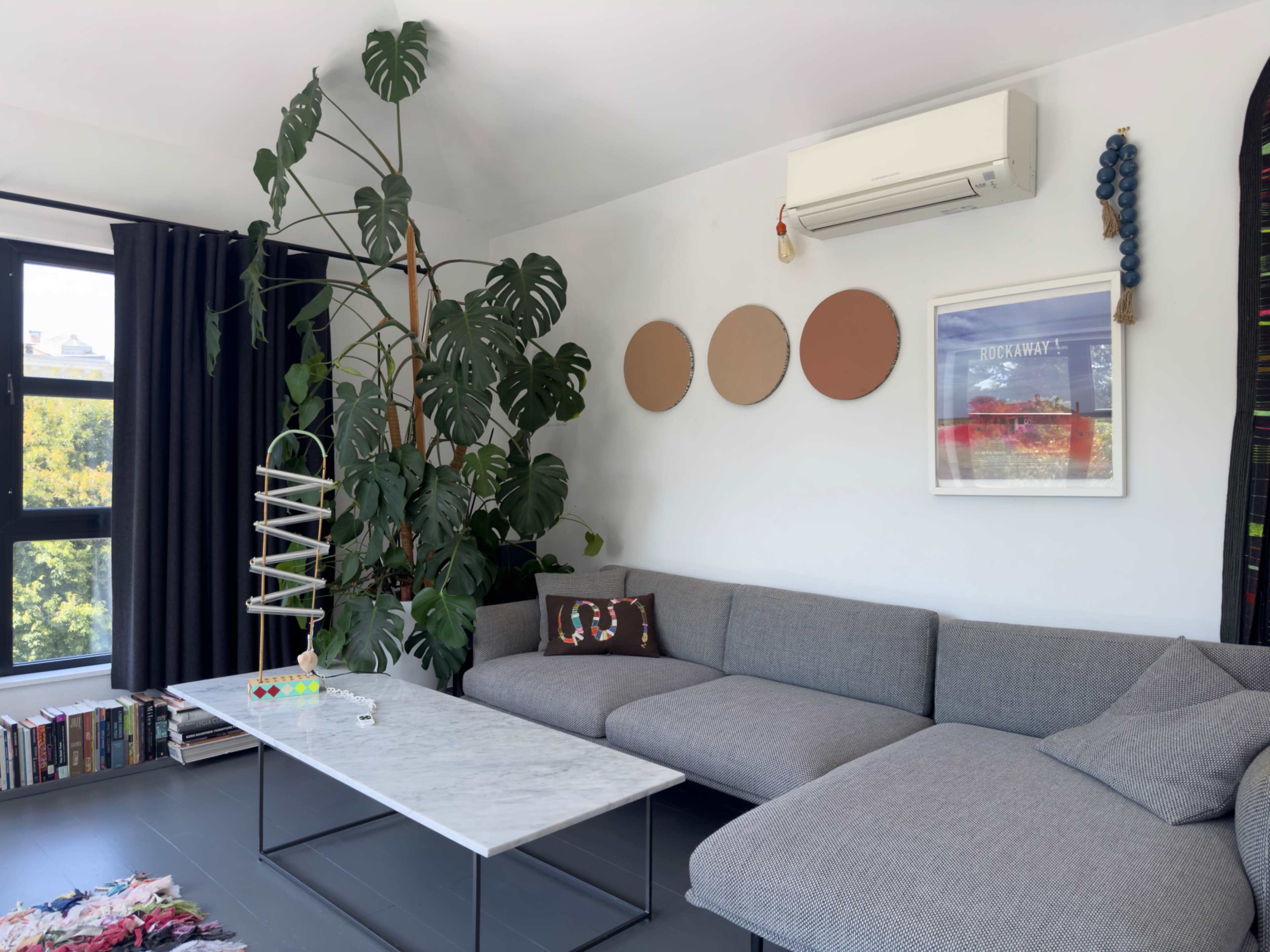 A modern living room features a gray sectional sofa, a marble coffee table, artistic wall decor, a large houseplant, and a bookshelf.