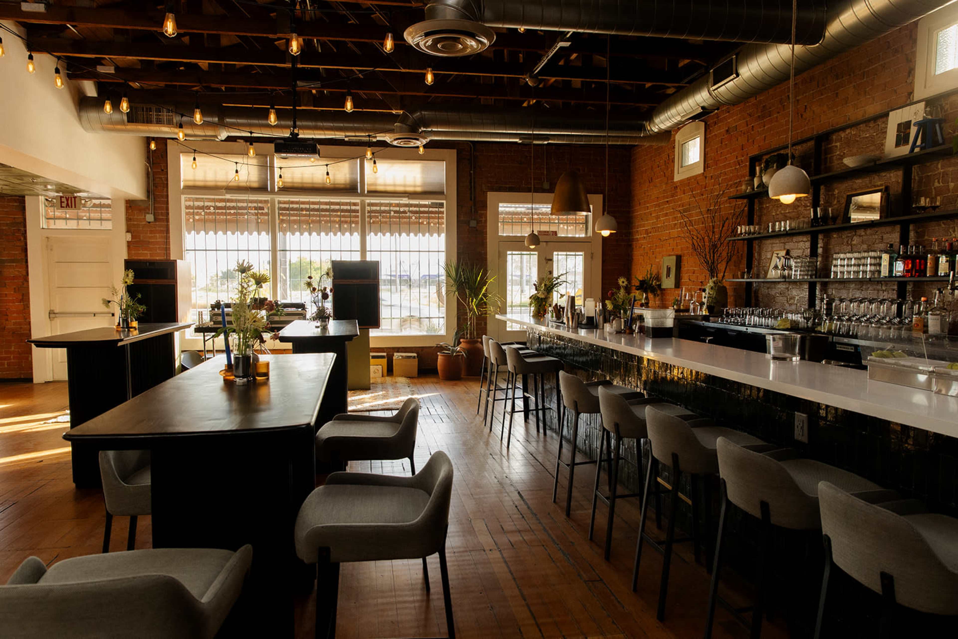 The image shows a modern bar and restaurant interior with wooden beams, brick walls, and a long counter featuring stools, surrounded by tables and chairs.