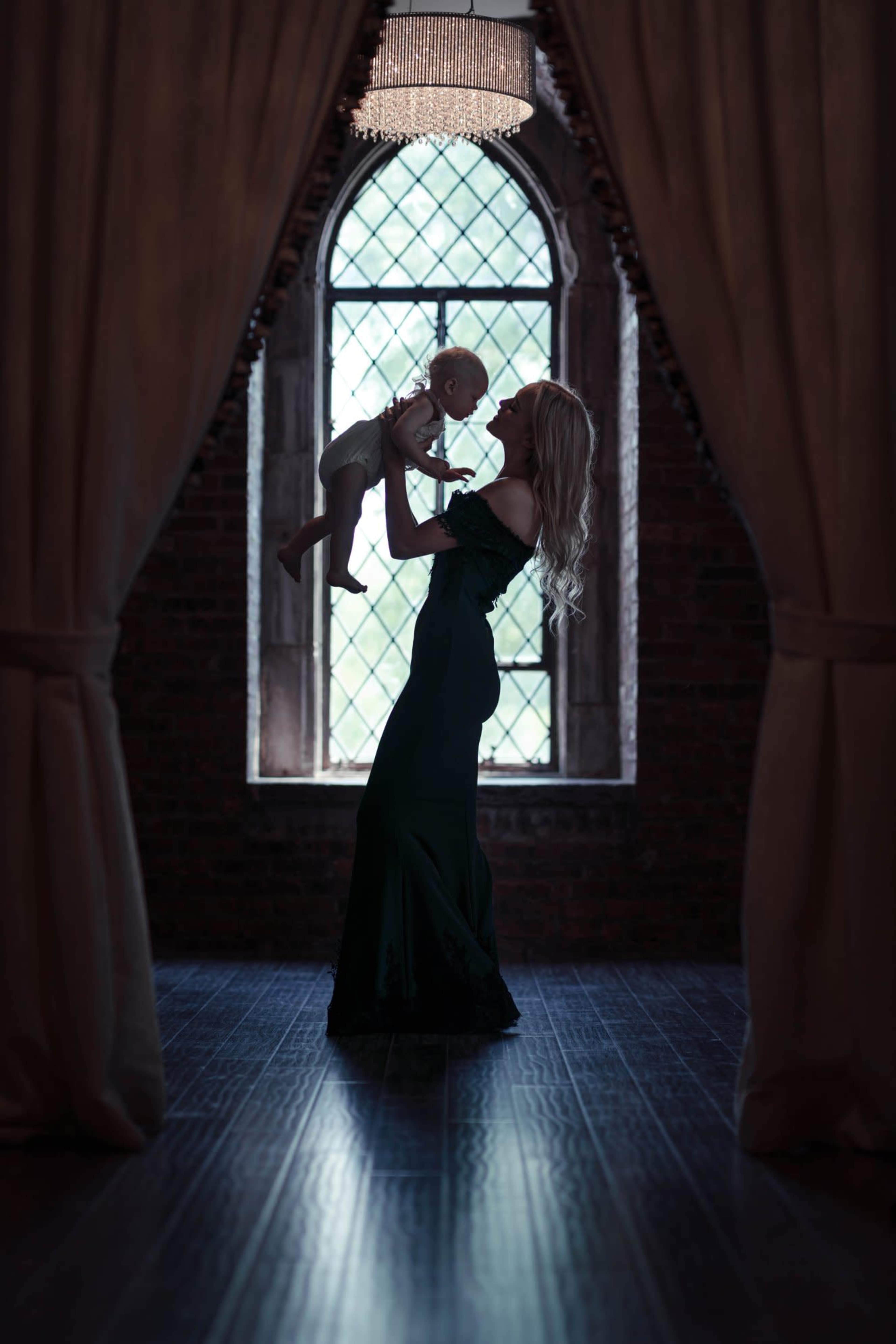 A woman in a dark dress is holding a baby in front of a large window with intricate glass patterns.