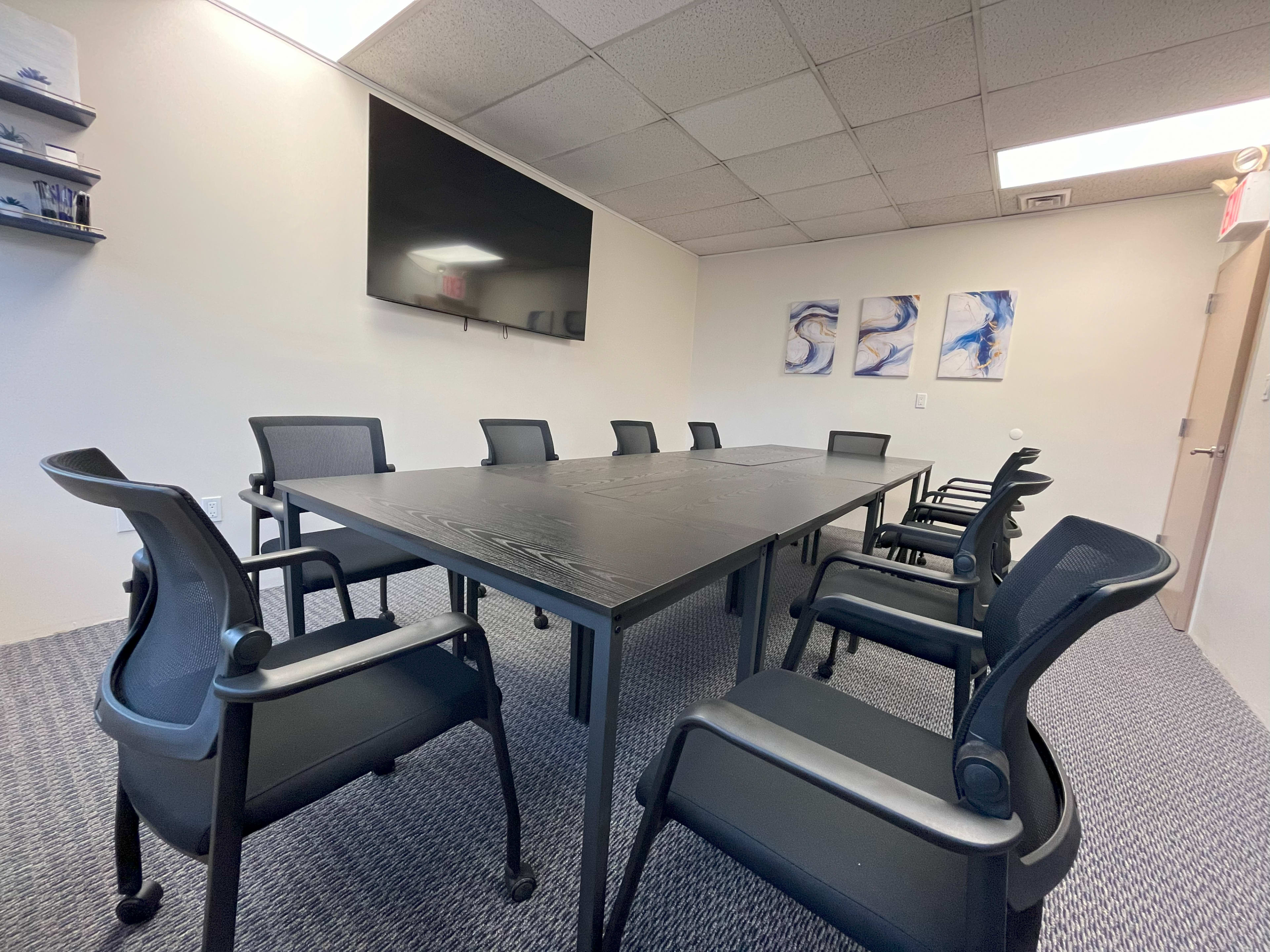 A conference room is set up with a large black table and eight black chairs, featuring a mounted television and three abstract paintings on the wall.