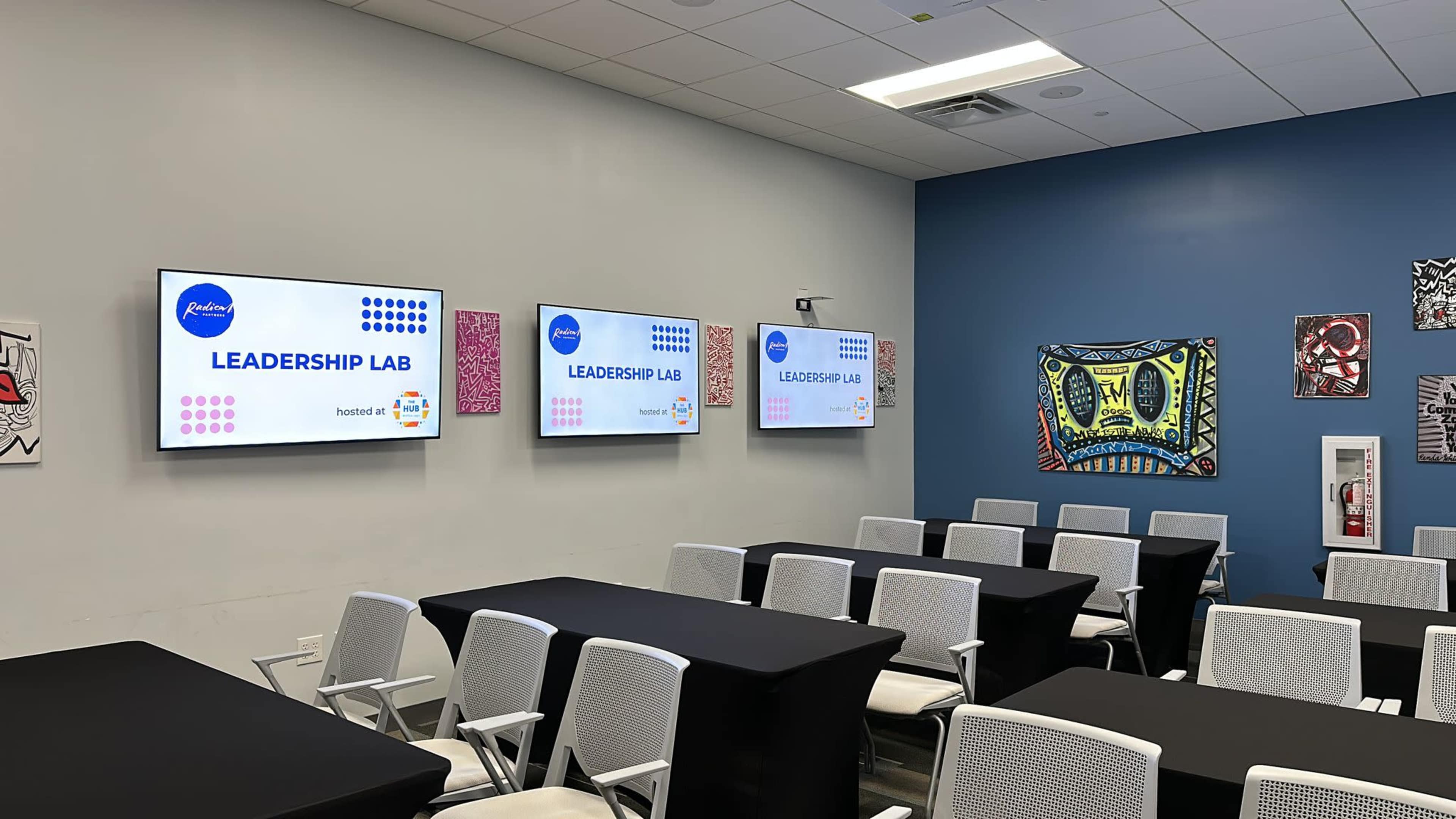 The image shows a classroom setup with black tables and three wall-mounted screens displaying the text "LEADERSHIP LAB" in a modern meeting space.