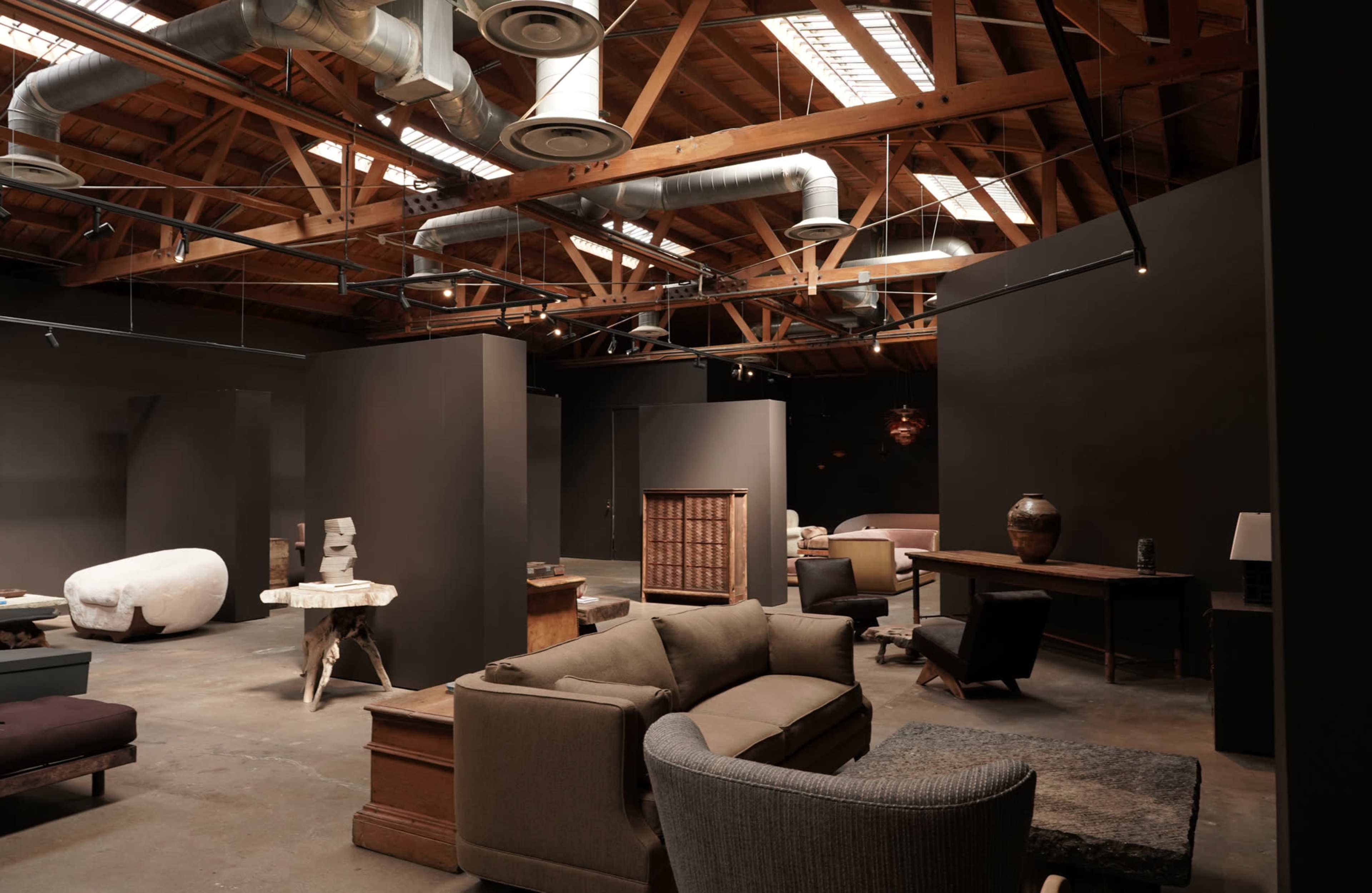 The image shows a spacious interior with industrial wooden beams, showcasing various modern furniture pieces arranged in distinct zones.
