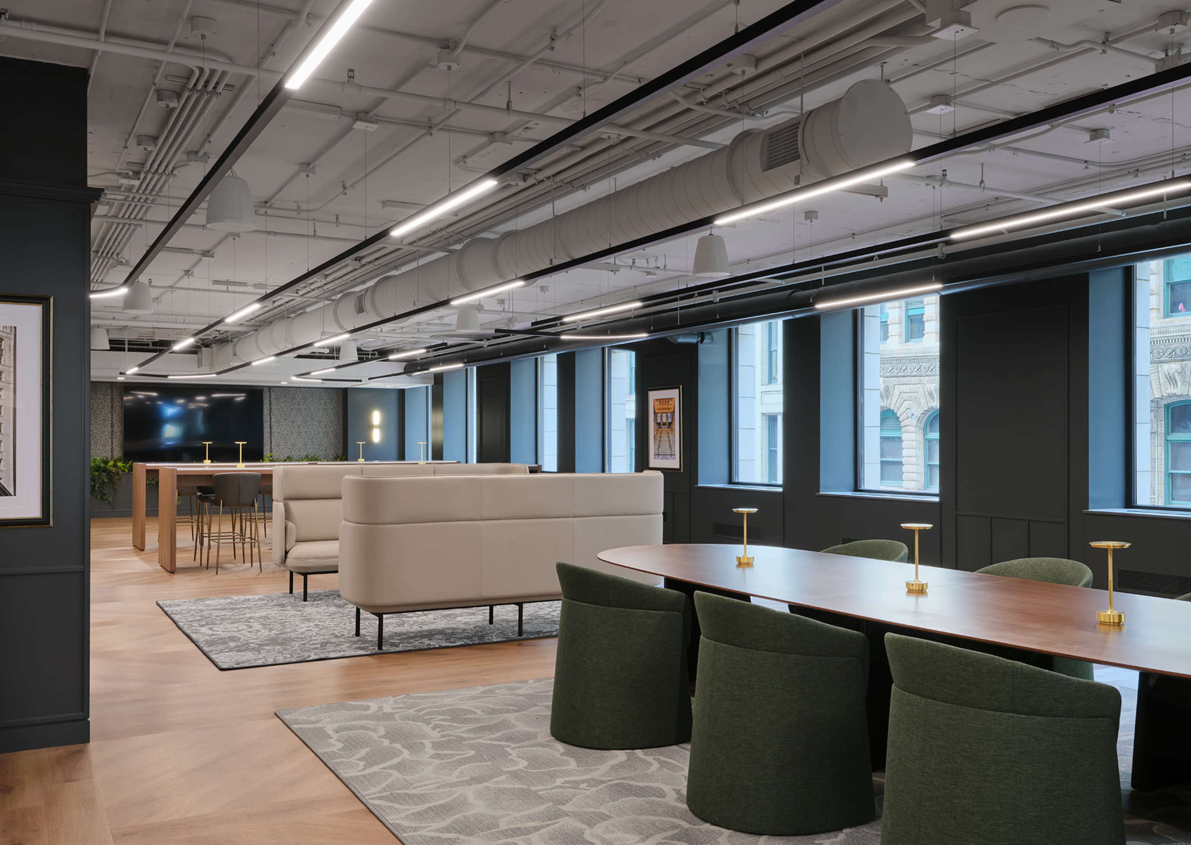 The image shows a modern office interior with a large conference table, green chairs, and various seating areas surrounded by large windows.