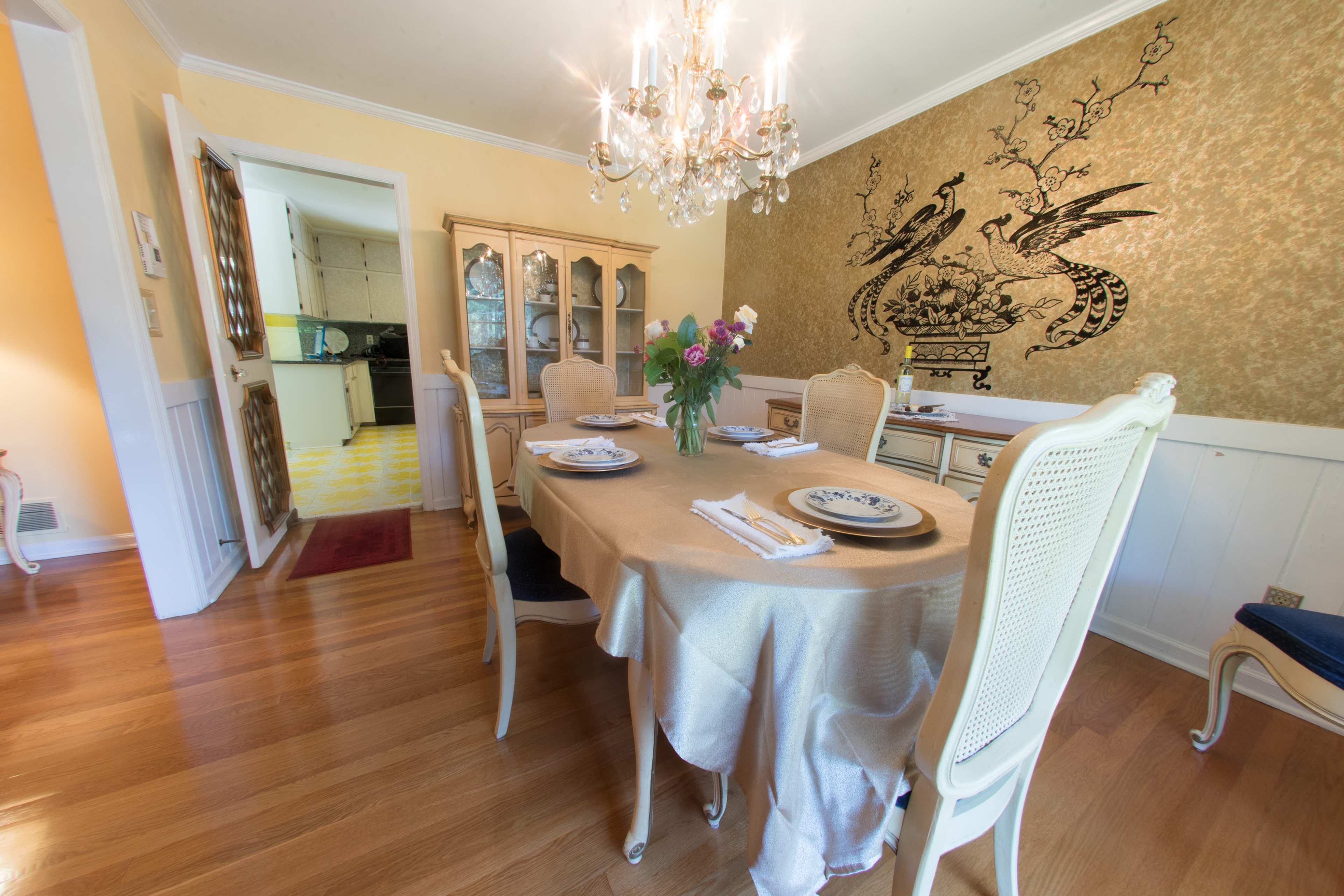 The dining room features a round table set with plates and a vase of flowers, with a chandelier above and a patterned wall across from the entrance.