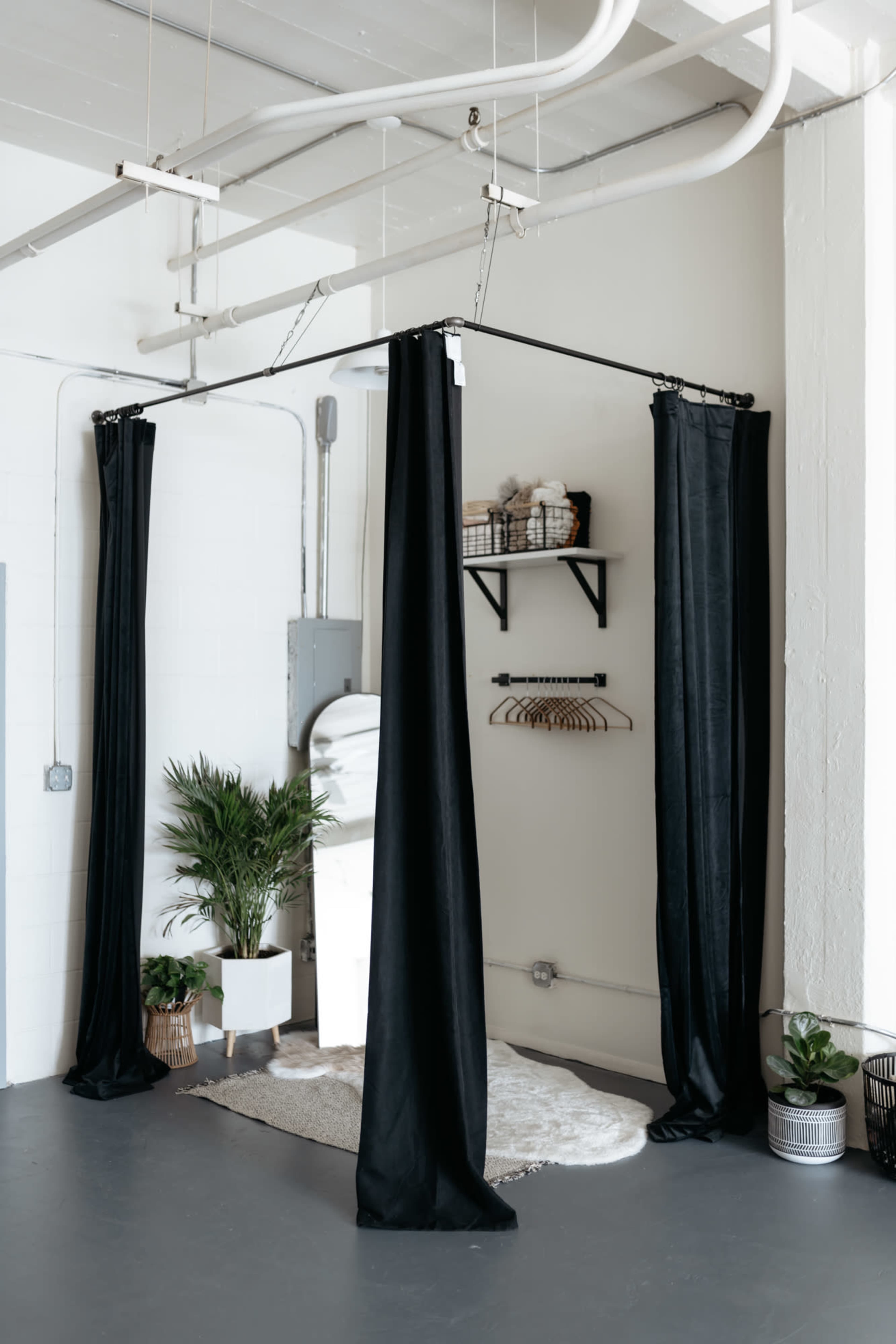 A minimalistic changing area is defined by black curtains, with a shelf holding storage bins and a plant beside a mirror.