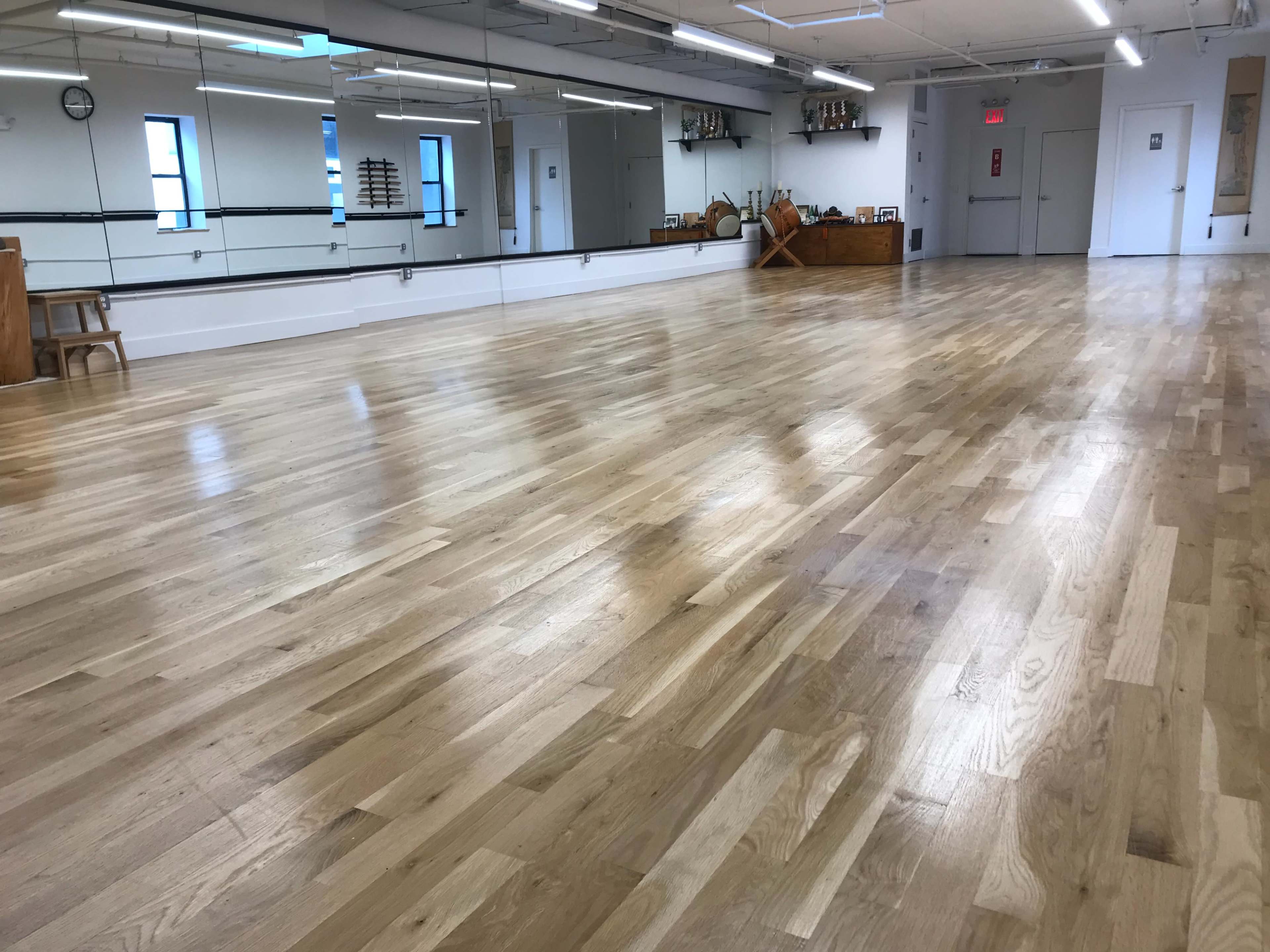 The image shows a spacious, empty dance studio with wooden flooring, large mirrors on one wall, and natural light coming through windows.