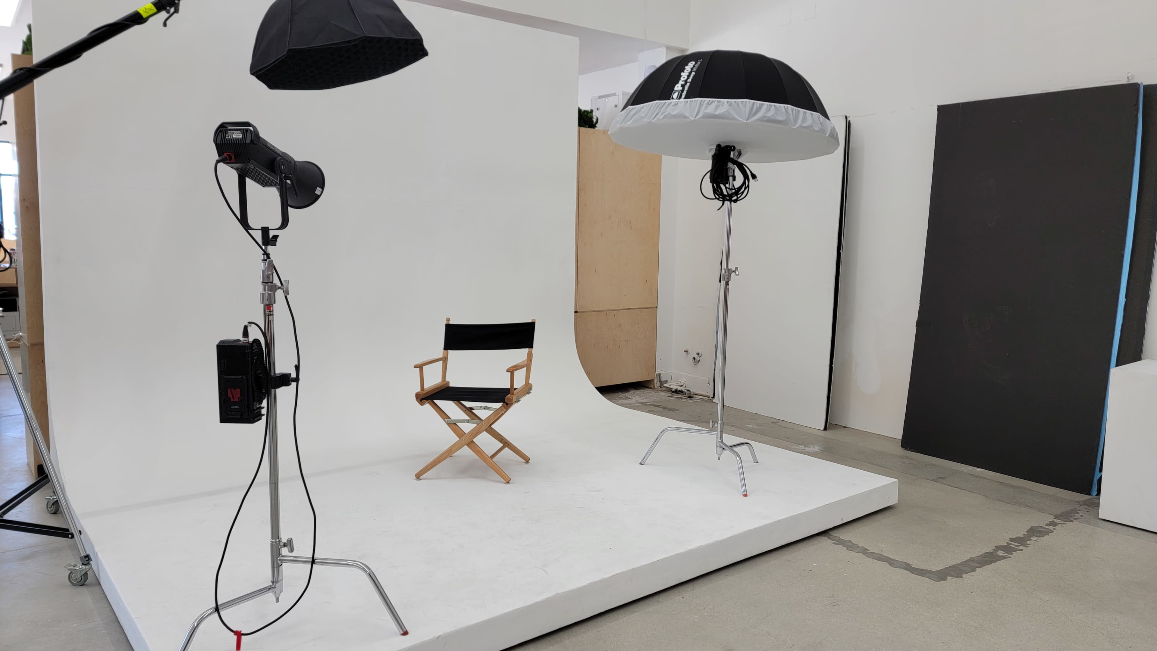 A photography studio setup features a director's chair on a white platform, flanked by two lighting umbrellas and a spotlight.