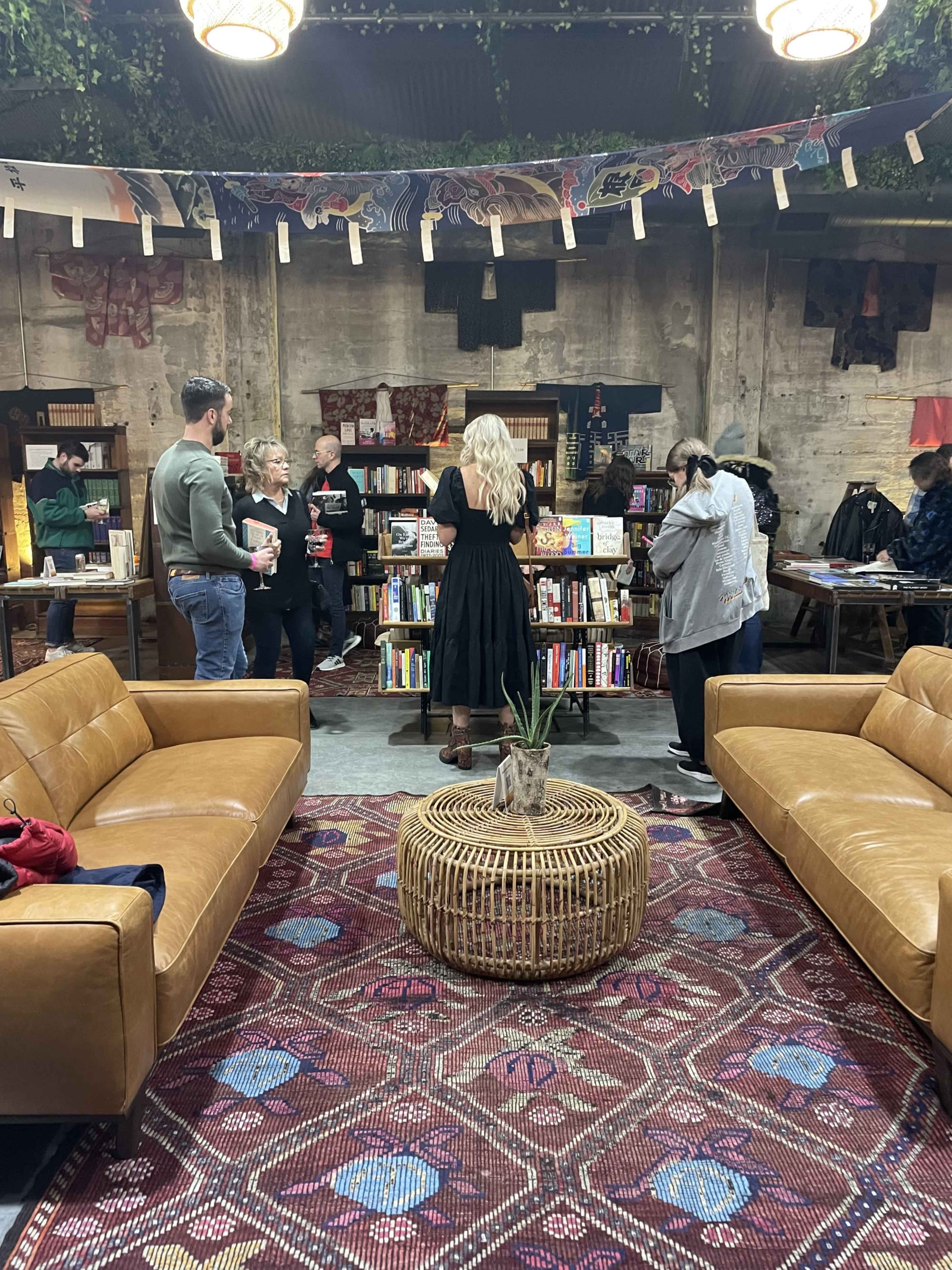 A group of people gathers in a cozy bookstore adorned with bookshelves and a patterned rug, while two leather sofas are arranged nearby.