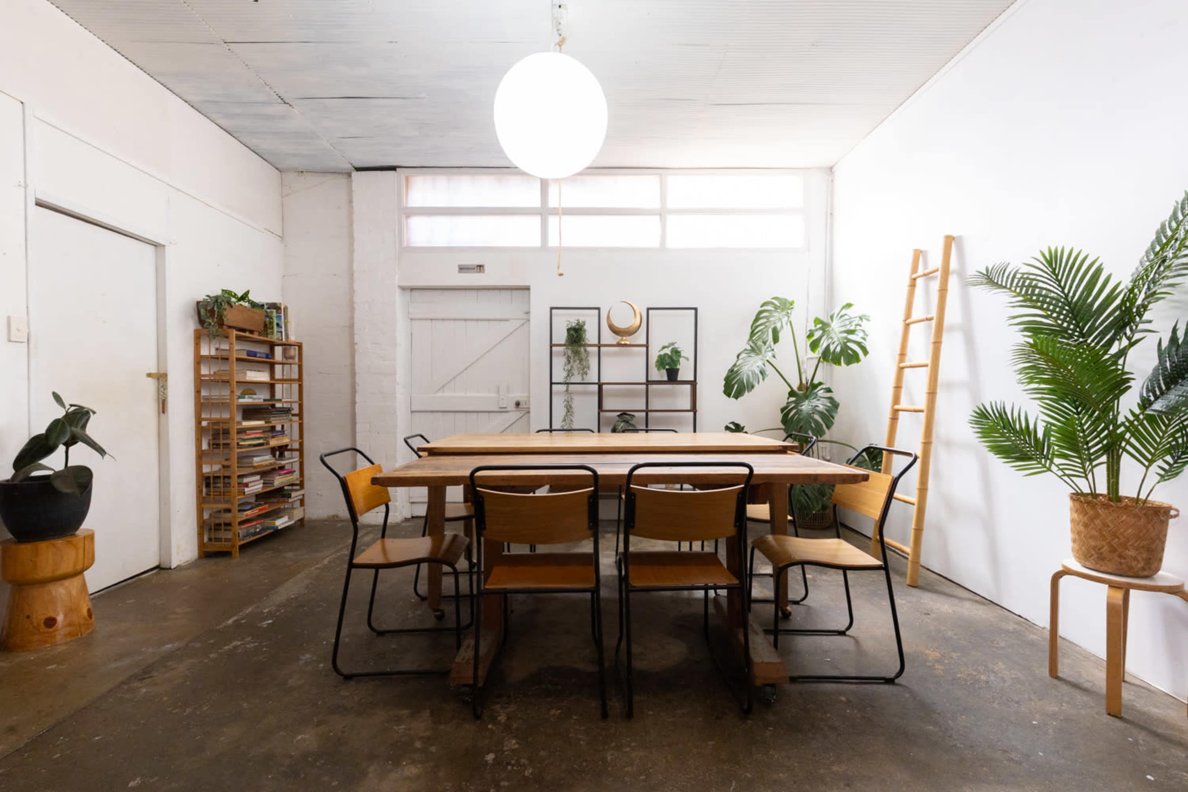 A spacious room features a large wooden table surrounded by chairs, with plants and bookshelves along the walls.
