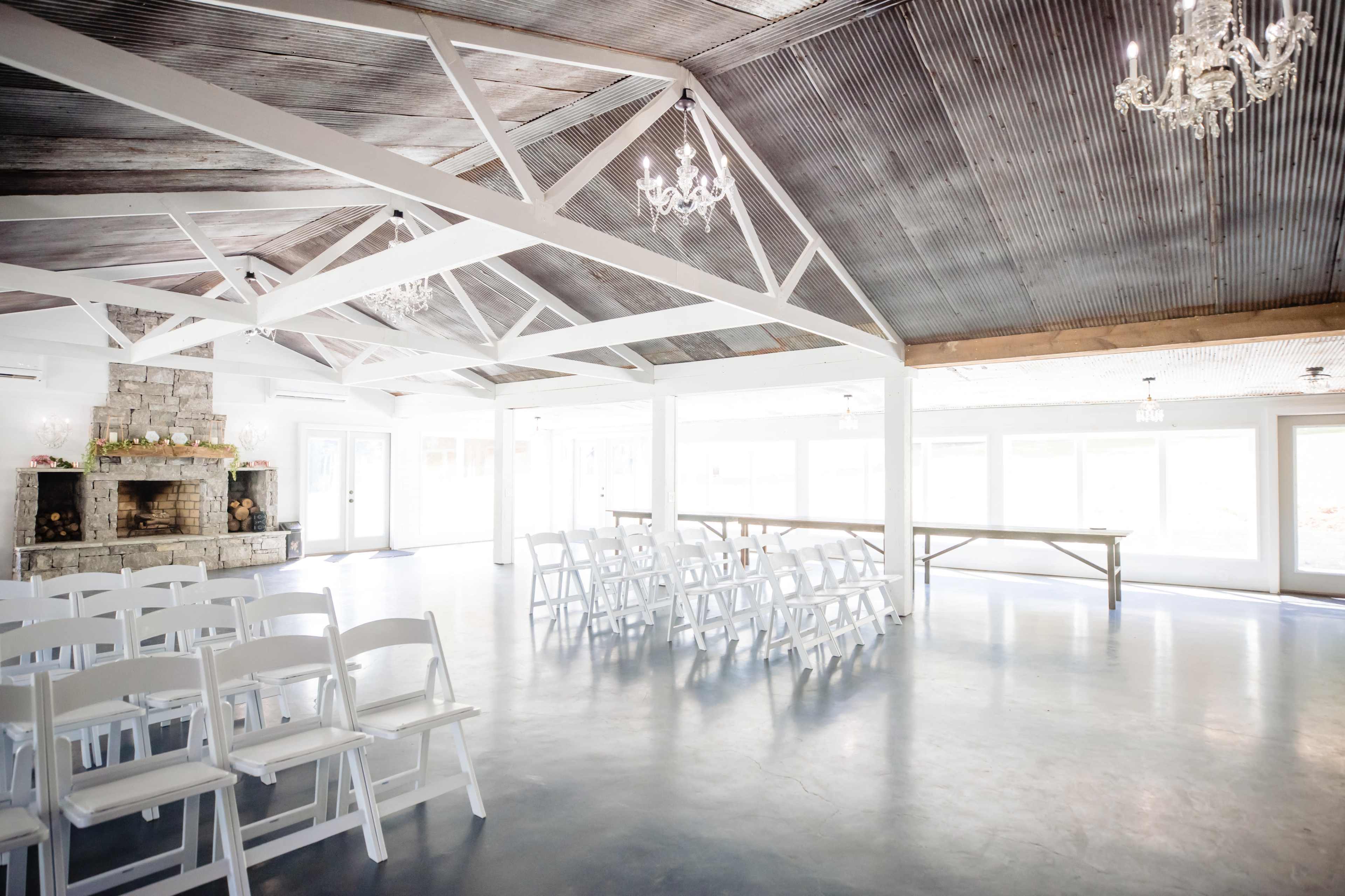 The interior of a spacious event venue features white folding chairs arranged facing a central area, with a stone fireplace and chandeliers enhancing the rustic ceiling.