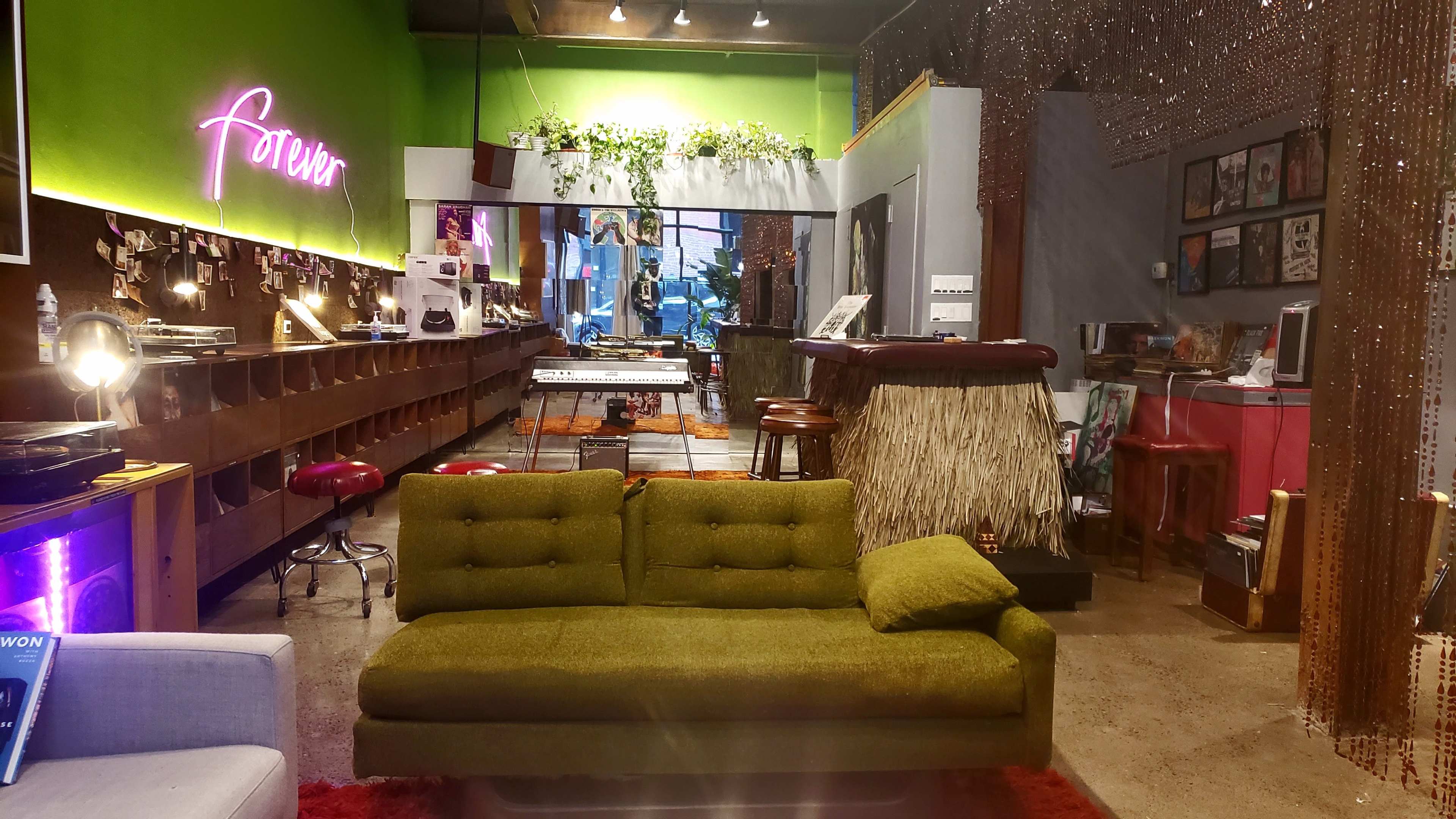 The scene features a retro-styled lounge area with a green sofa in the foreground, surrounded by a vibrant, eclectic space that includes a bar, musical instruments, and decorative plants.